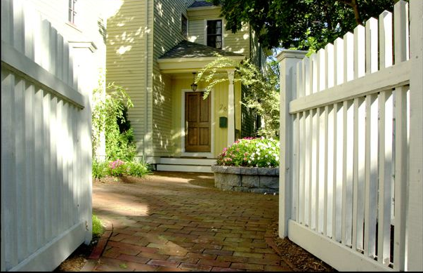Open white picket gate leading to a yellow house with a brick walkway and flowerbeds.
