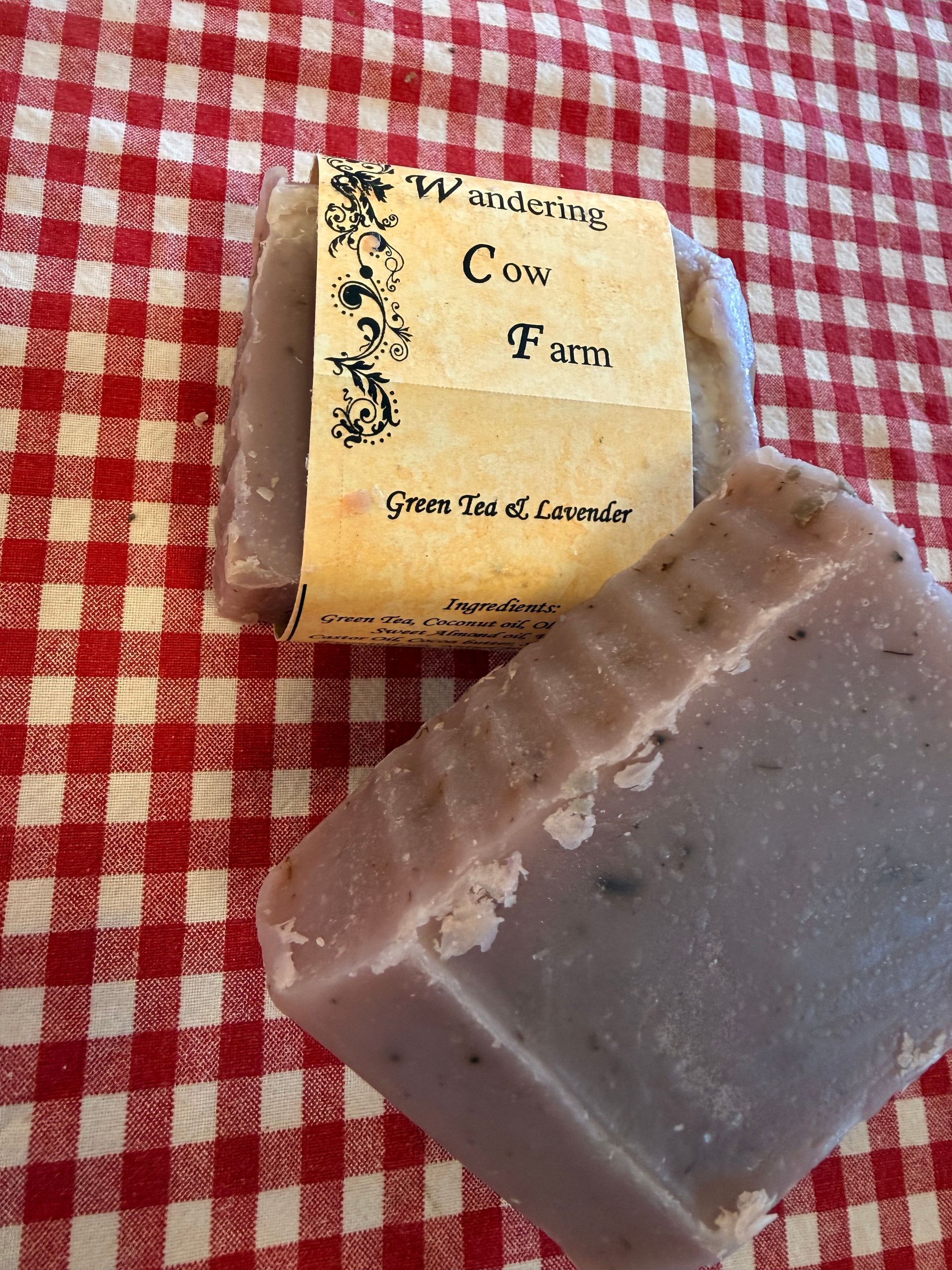 Two handmade soap bars on a red-and-white checkered cloth, one wrapped with a cream label.