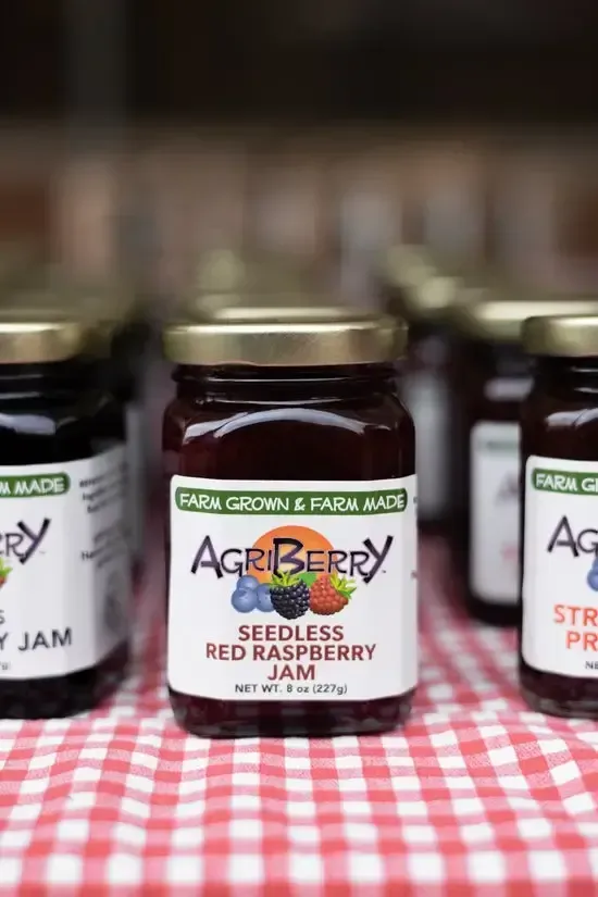 Several jars of AgriBerry jam with gold lids sit on a red and white checkered tablecloth.
