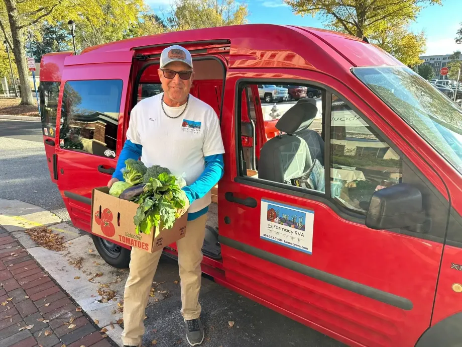 A person standing by an open red delivery van, holding a cardboard box filled with fresh leafy green vegetables.