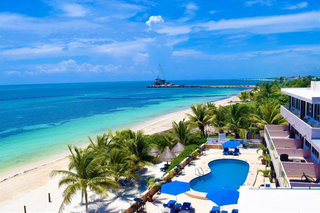 Beach resort with pool, ocean, white sand, palm trees, and blue umbrellas.