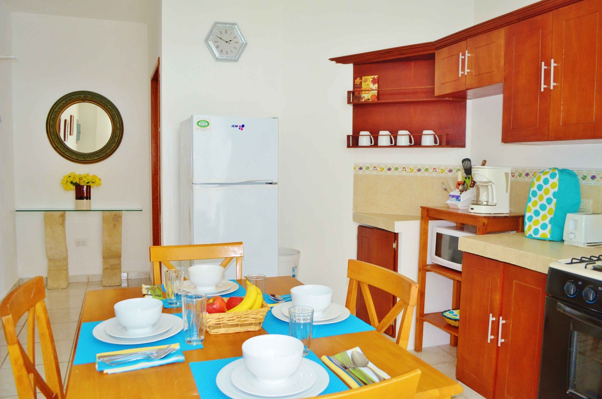 Cocina con mesa de comedor, armarios de madera, refrigerador blanco y vajilla para cuatro personas.