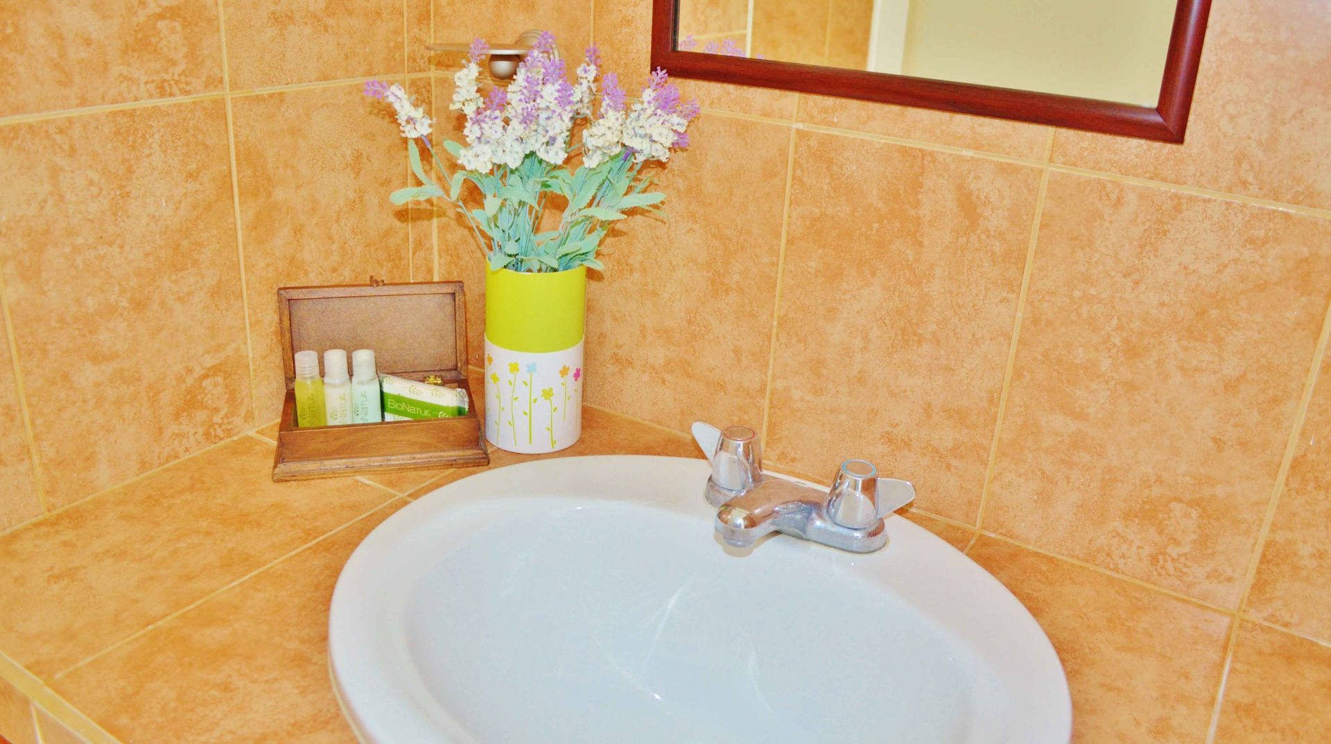 Bathroom sink with a floral arrangement, toiletries, and a mirror against orange-tiled walls.