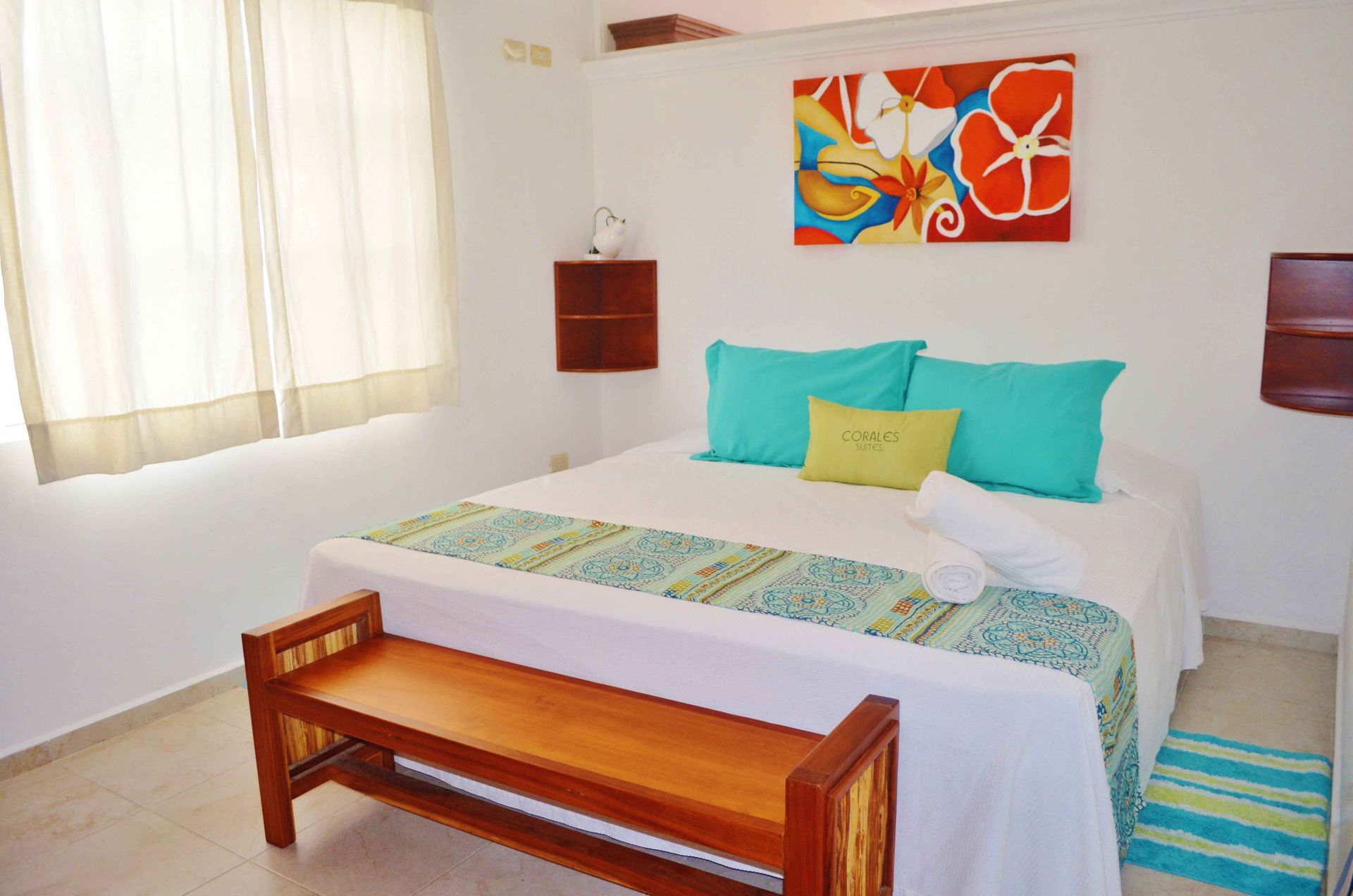 Dormitorio con cama blanca, almohadas turquesa, banco y un cuadro colorido.