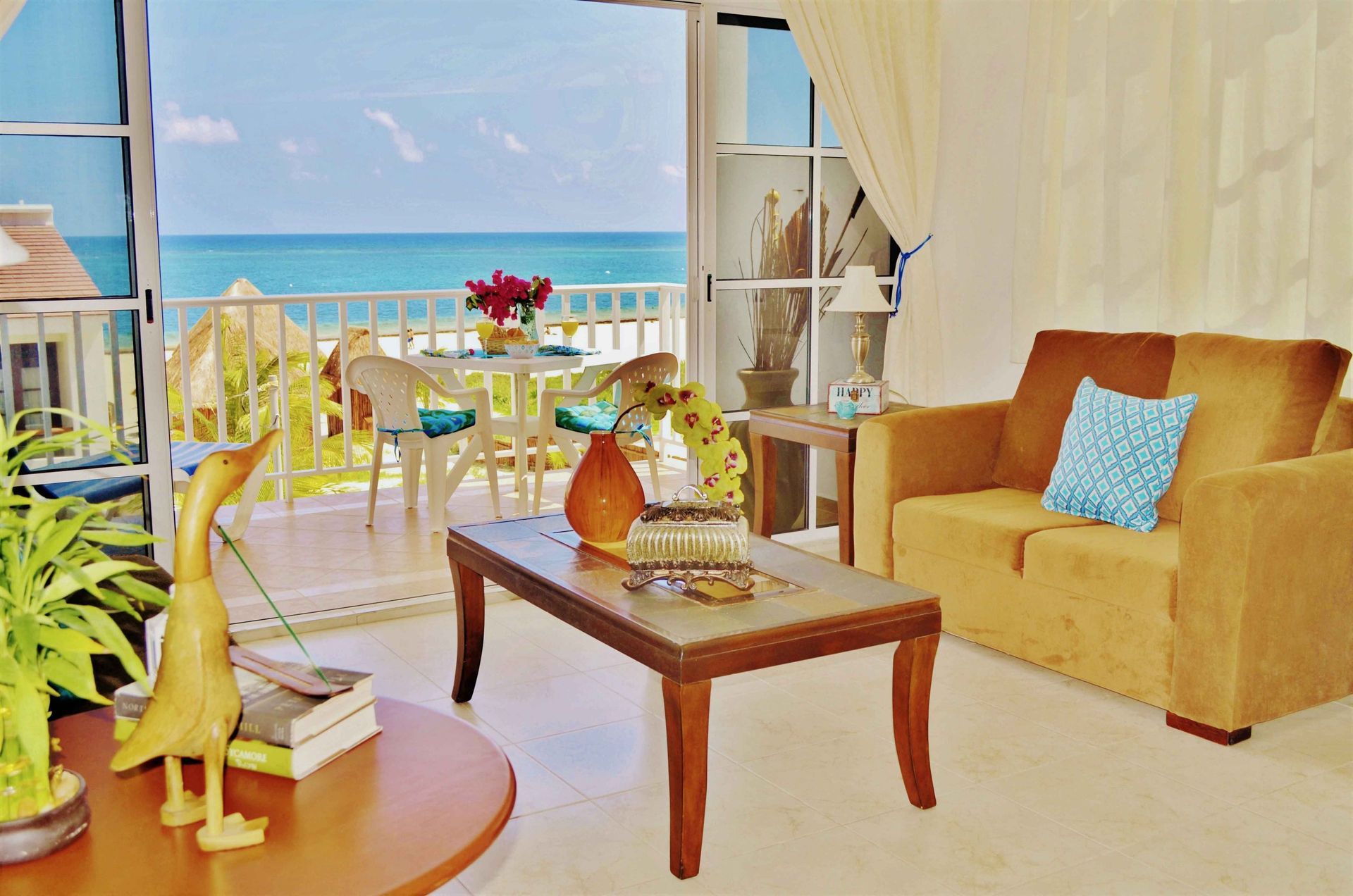 Sala de estar con vista al mar, sofá amarillo, mesa de centro y mesa de balcón con flores.