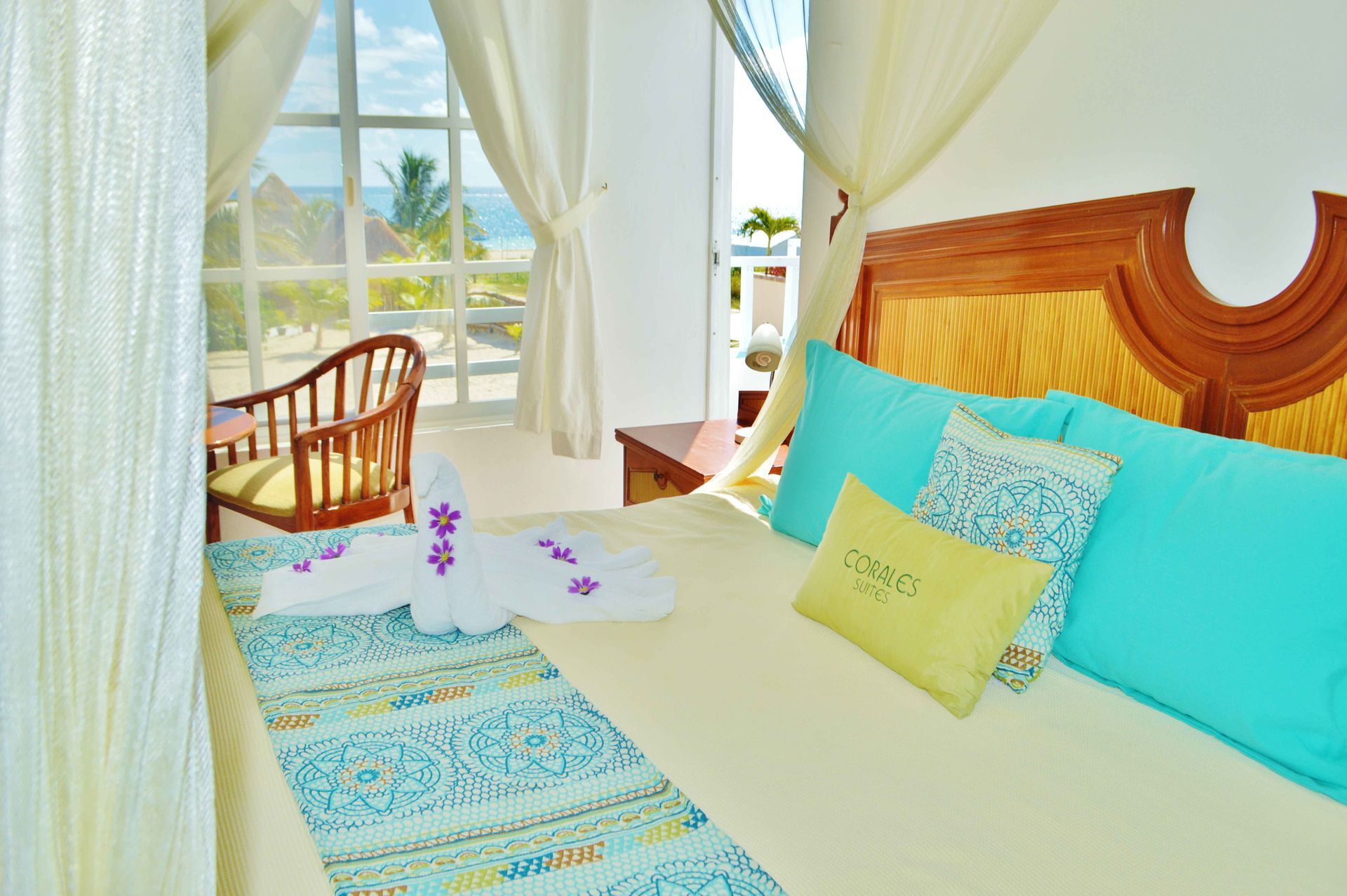 Dormitorio con vistas al mar, cama grande con almohadas turquesa, cortinas blancas y una silla de madera.