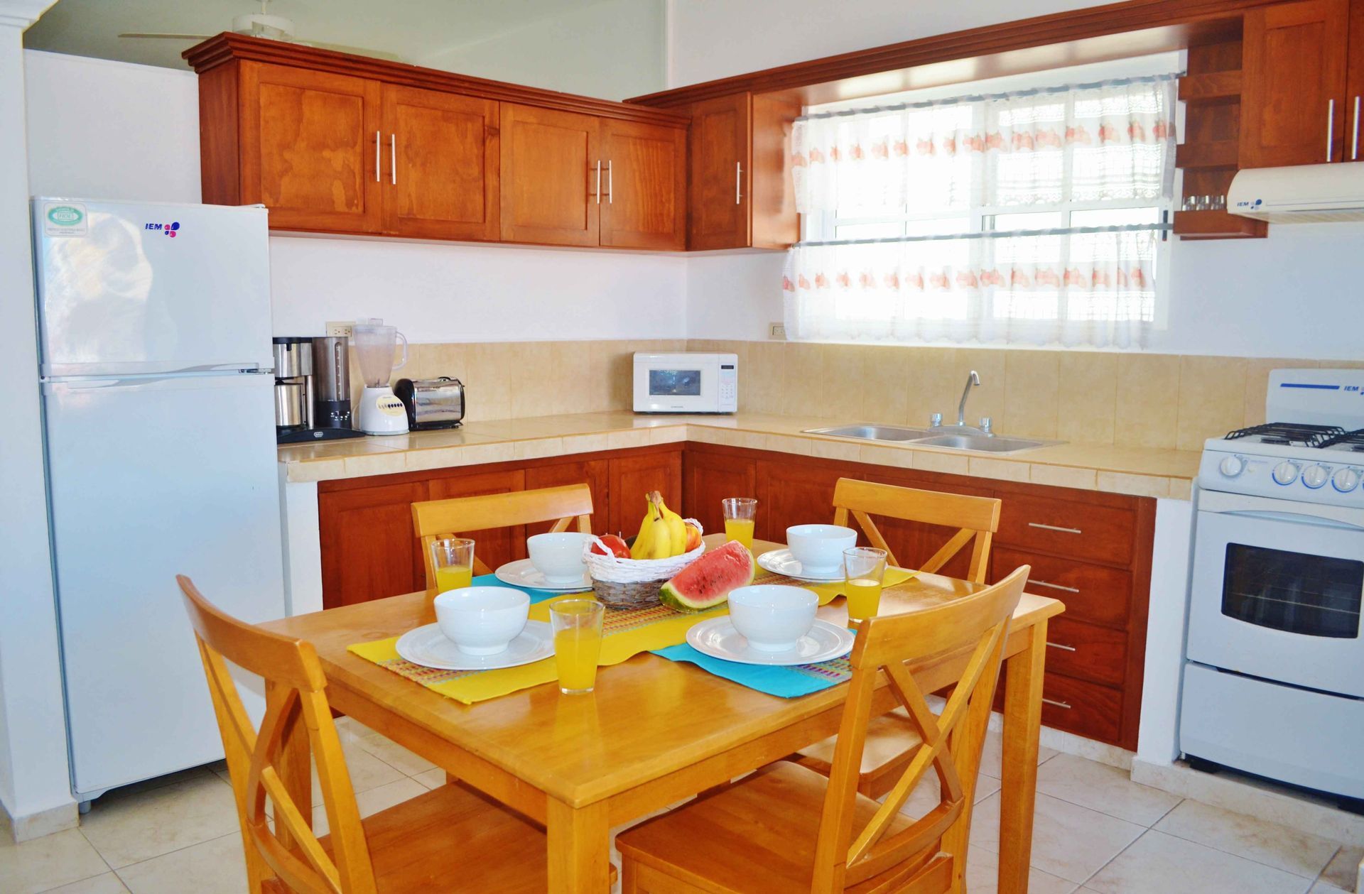 Cocina con armarios de madera, electrodomésticos blancos, mesa de comedor puesta para desayunar con fruta y zumo de naranja.