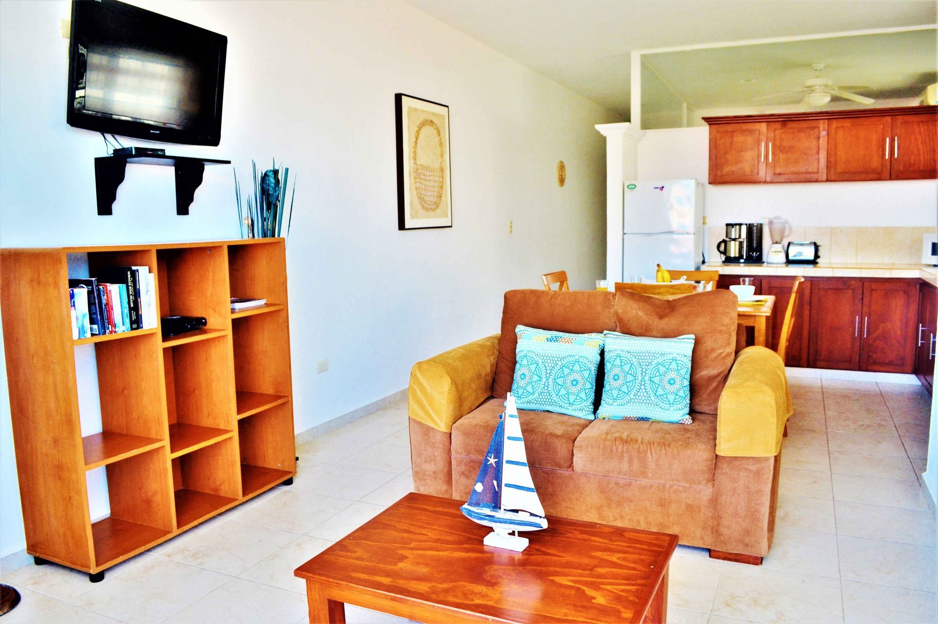 Sala de estar con sofá marrón, estantería y televisor. Cocina al fondo.