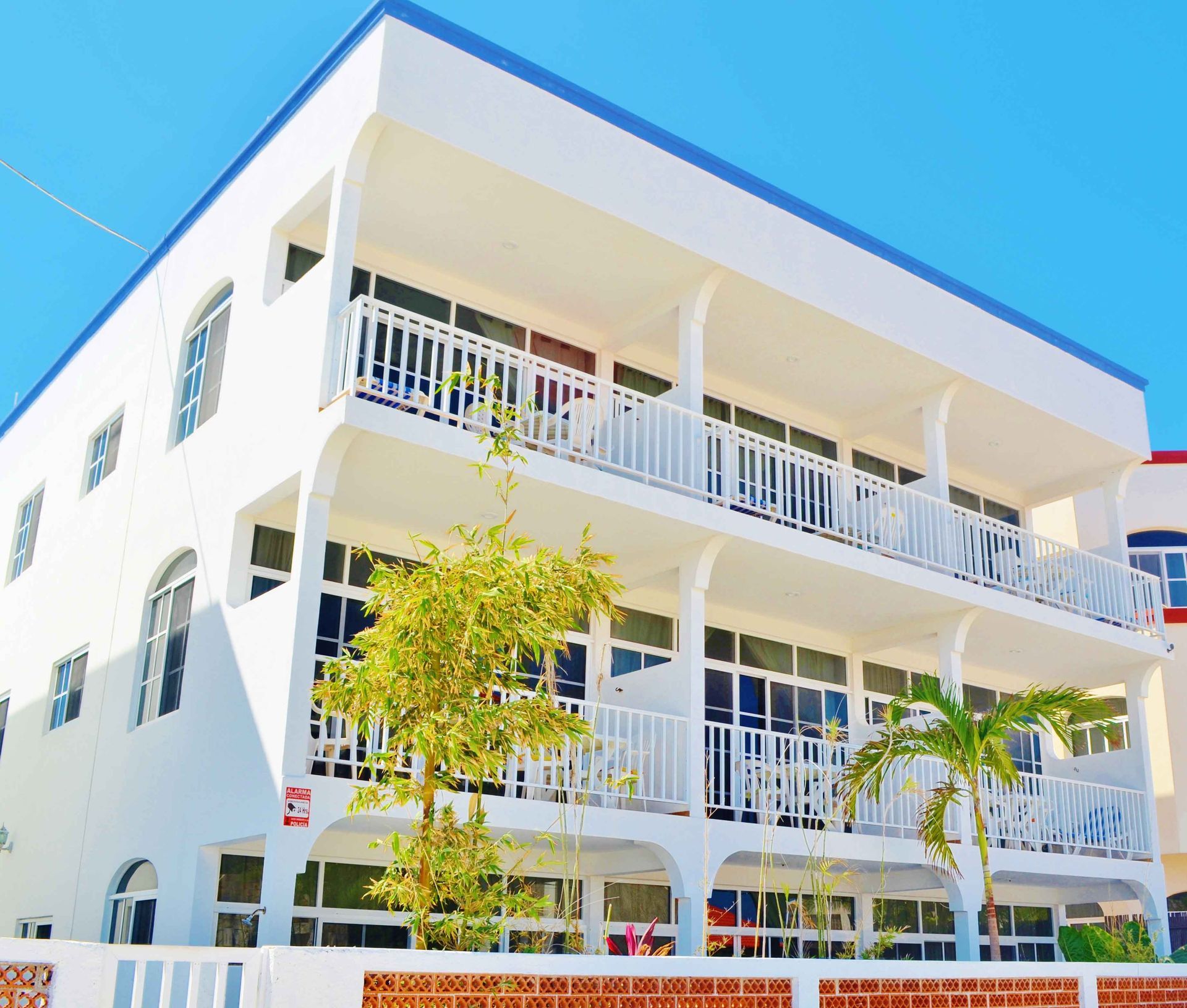 White three-story building with balconies, blue trim, and palm trees. Sunny day.