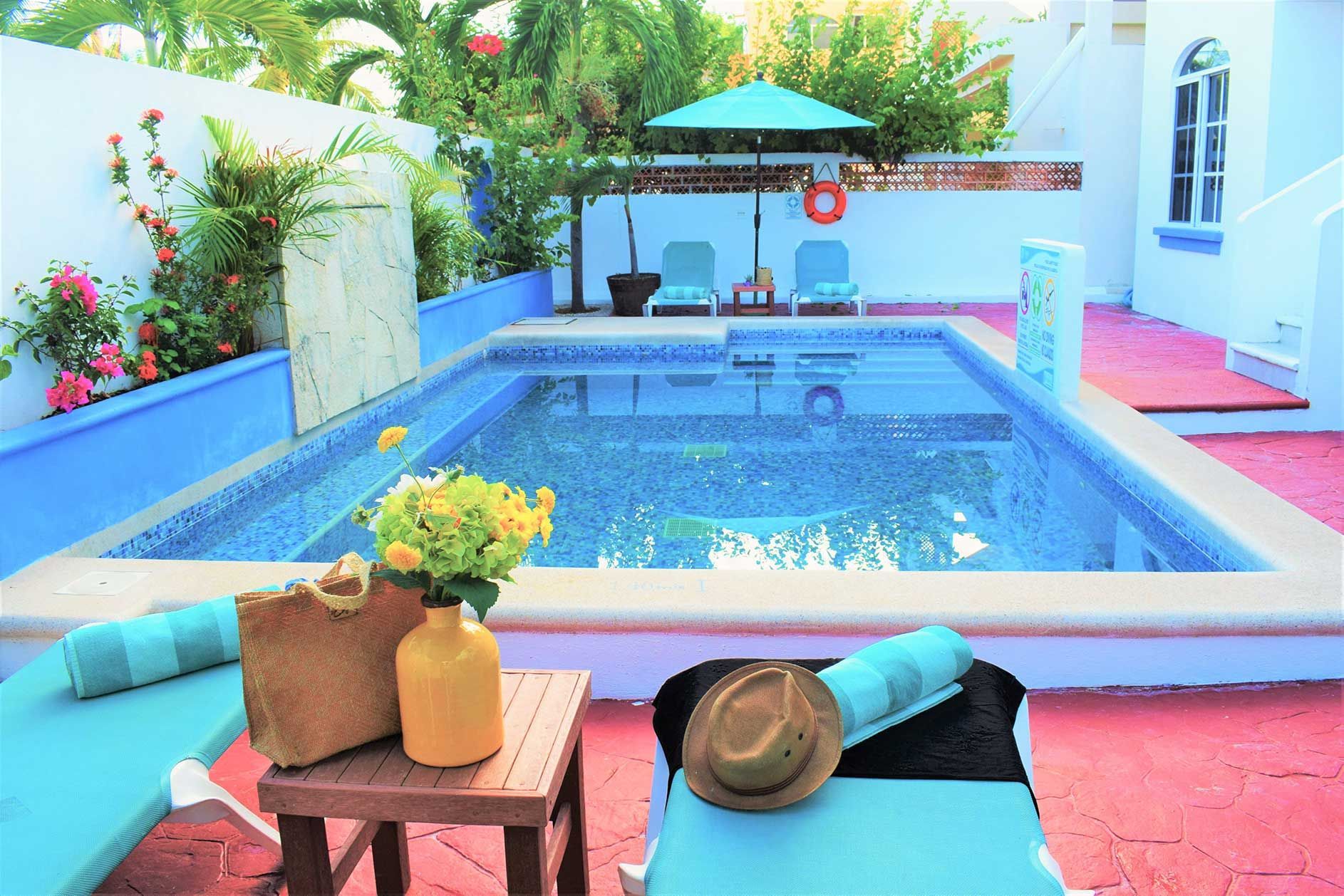 Poolside scene: pool, lounge chairs with towels, parasol, yellow vase with flowers.