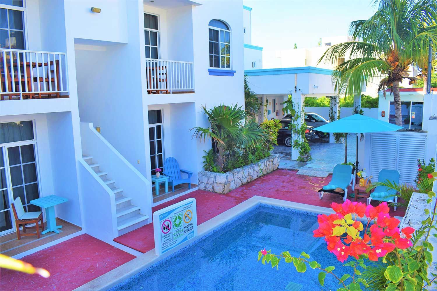 White building with a pool and lounge chairs. Palm trees, blue umbrella, red brick patio.
