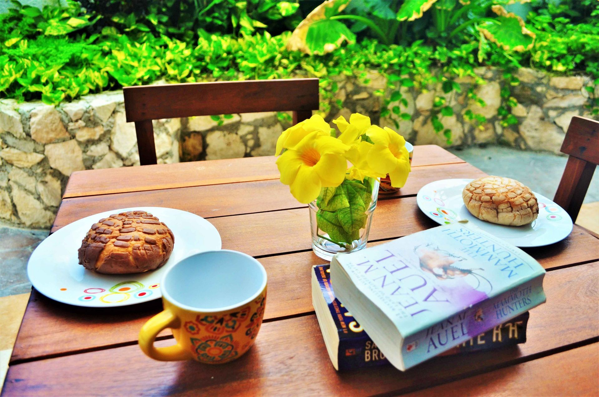 Wooden table with breakfast, yellow flowers, and a book on an outdoor patio.