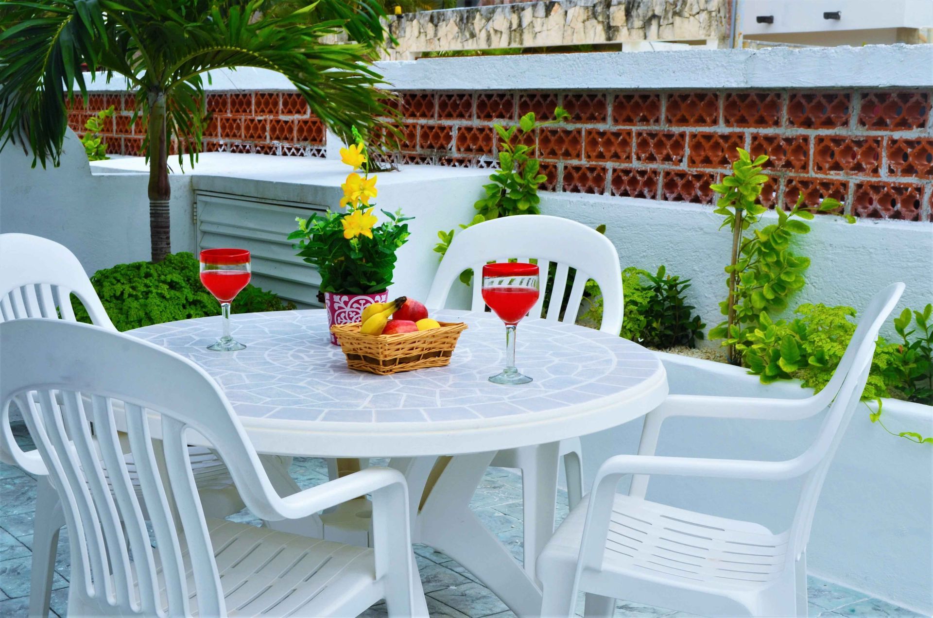 White plastic table and chairs on a patio with drinks, fruit, and plants.