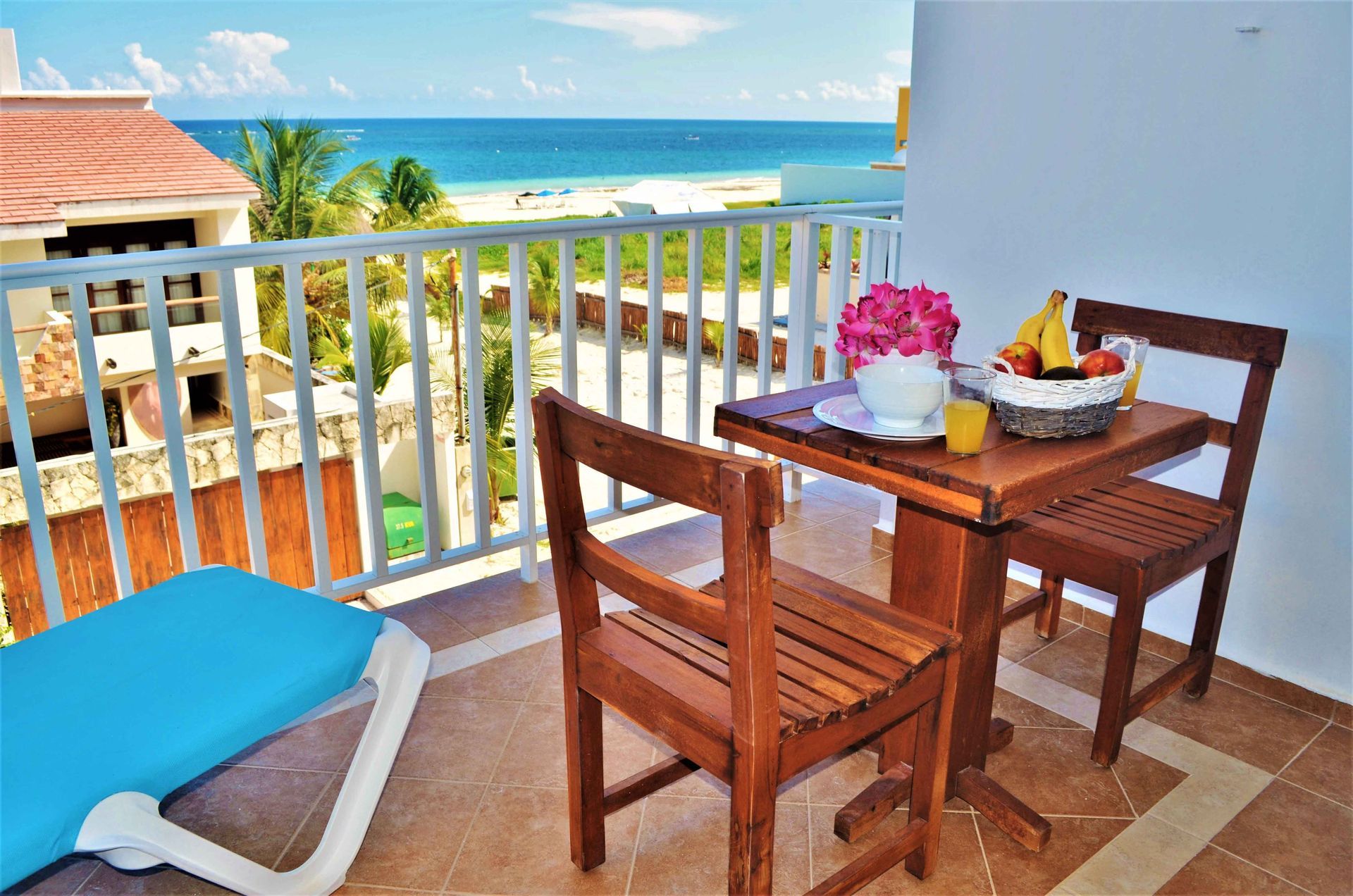 Balcón con mesa puesta para desayunar; vista al mar. Incluye cesta de frutas, jugo, flores y tumbona.