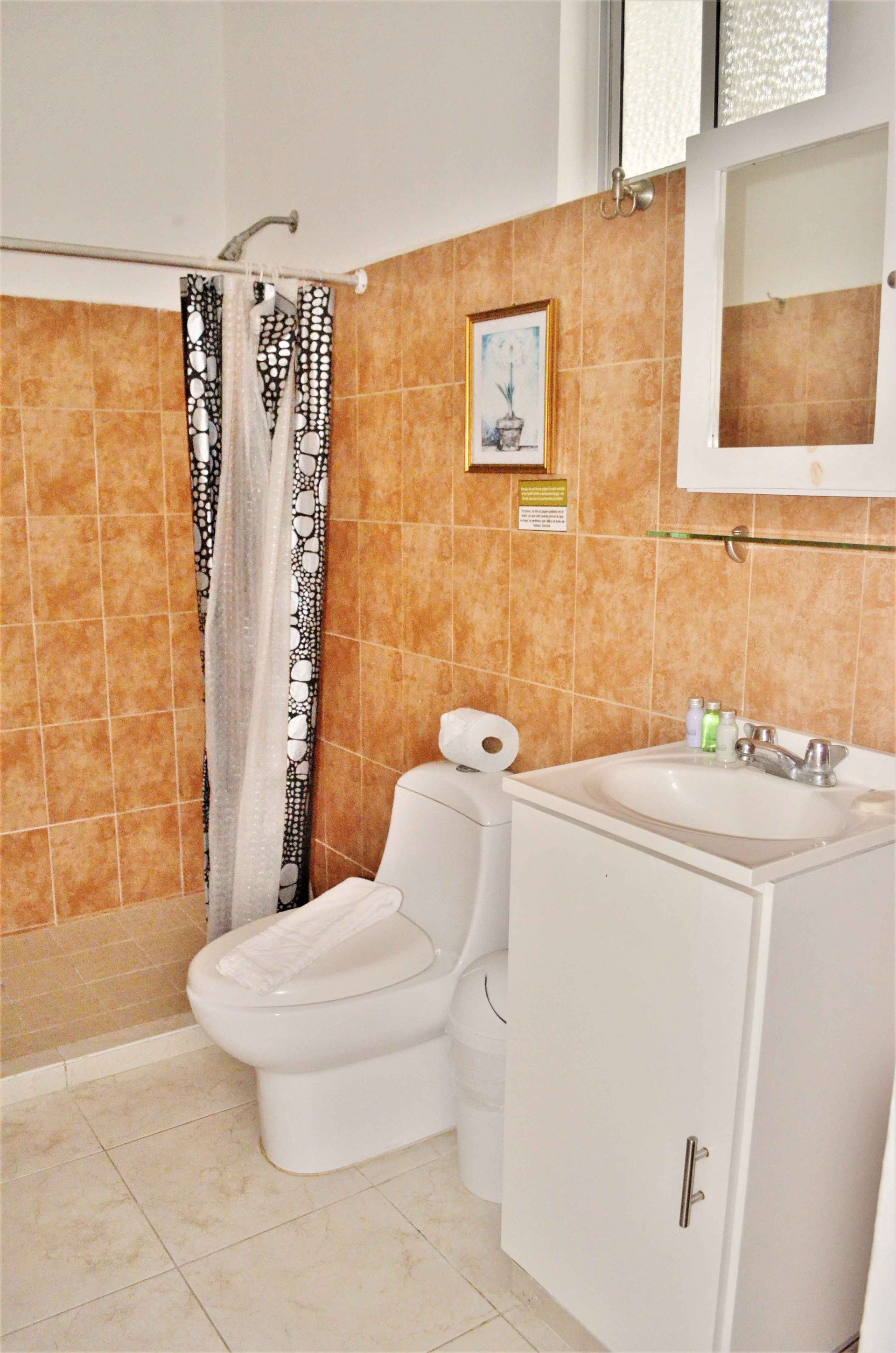Baño con inodoro, lavabo y ducha blancos con cortina blanca y negra; paredes alicatadas de color naranja.