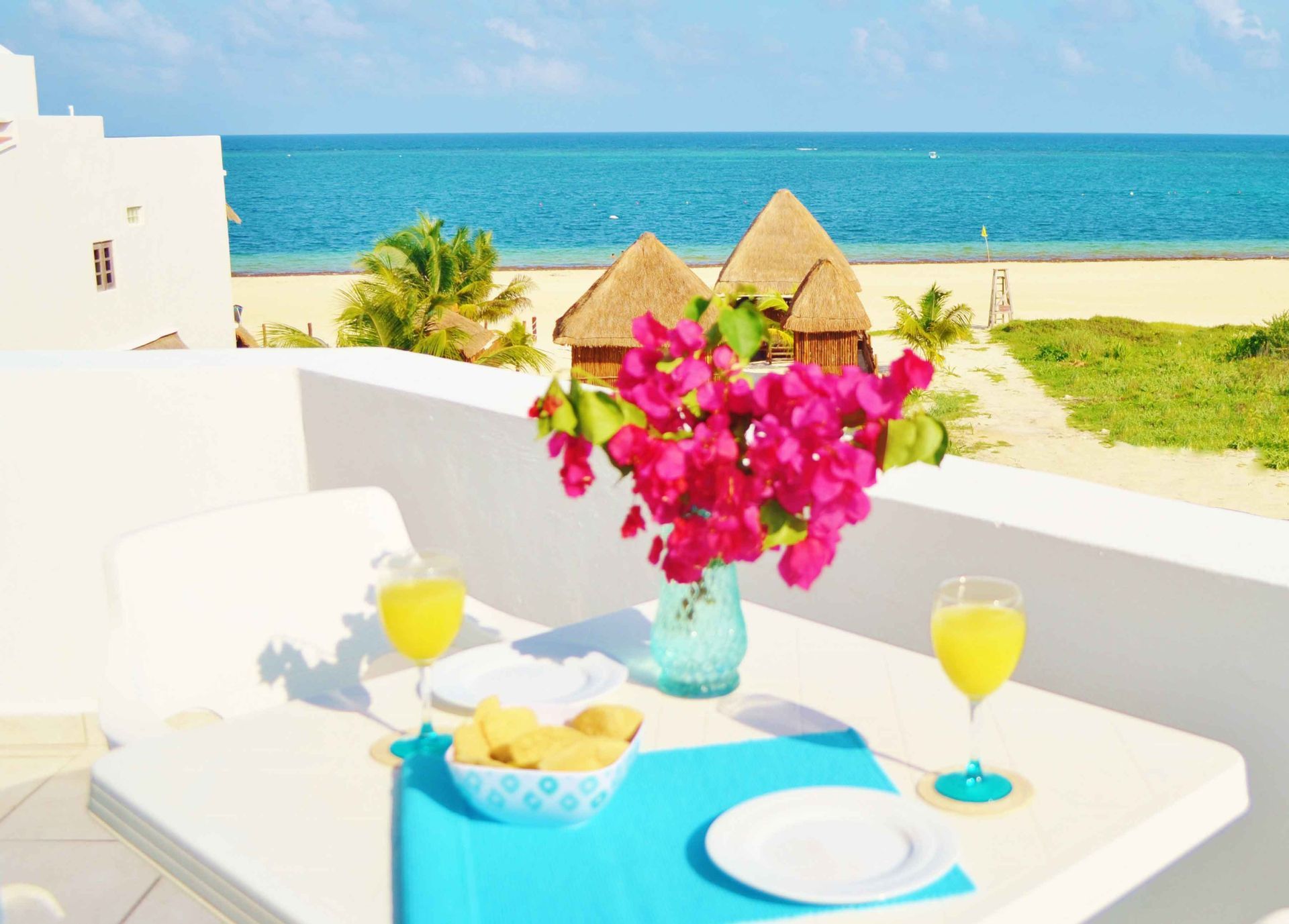 Mesa puesta para dos con vista al mar: bebidas, flores, aperitivos y detalles en color turquesa.