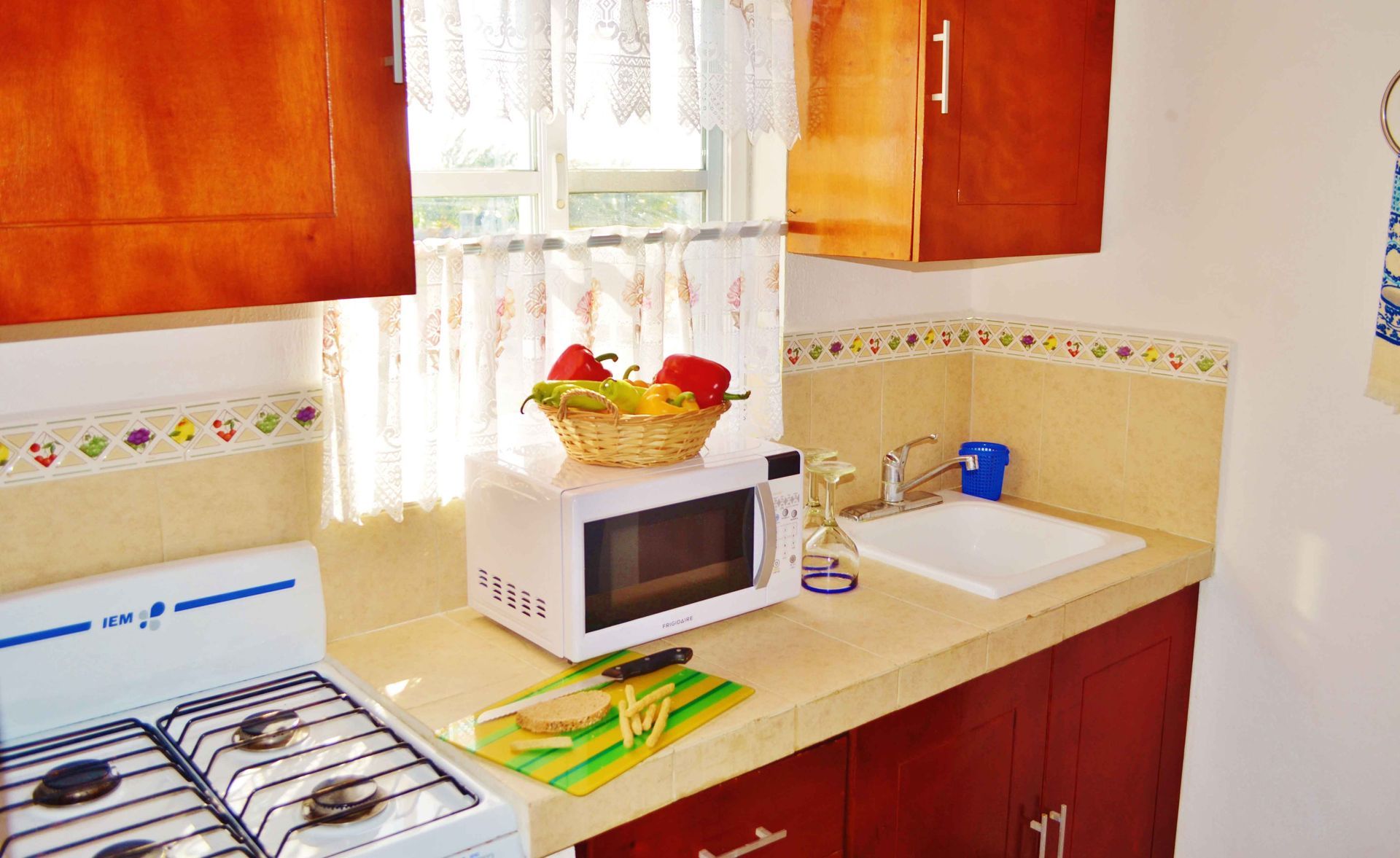 Pequeña cocina con armarios de madera, microondas, fregadero y fogones. Una cesta de fruta descansa sobre el microondas.