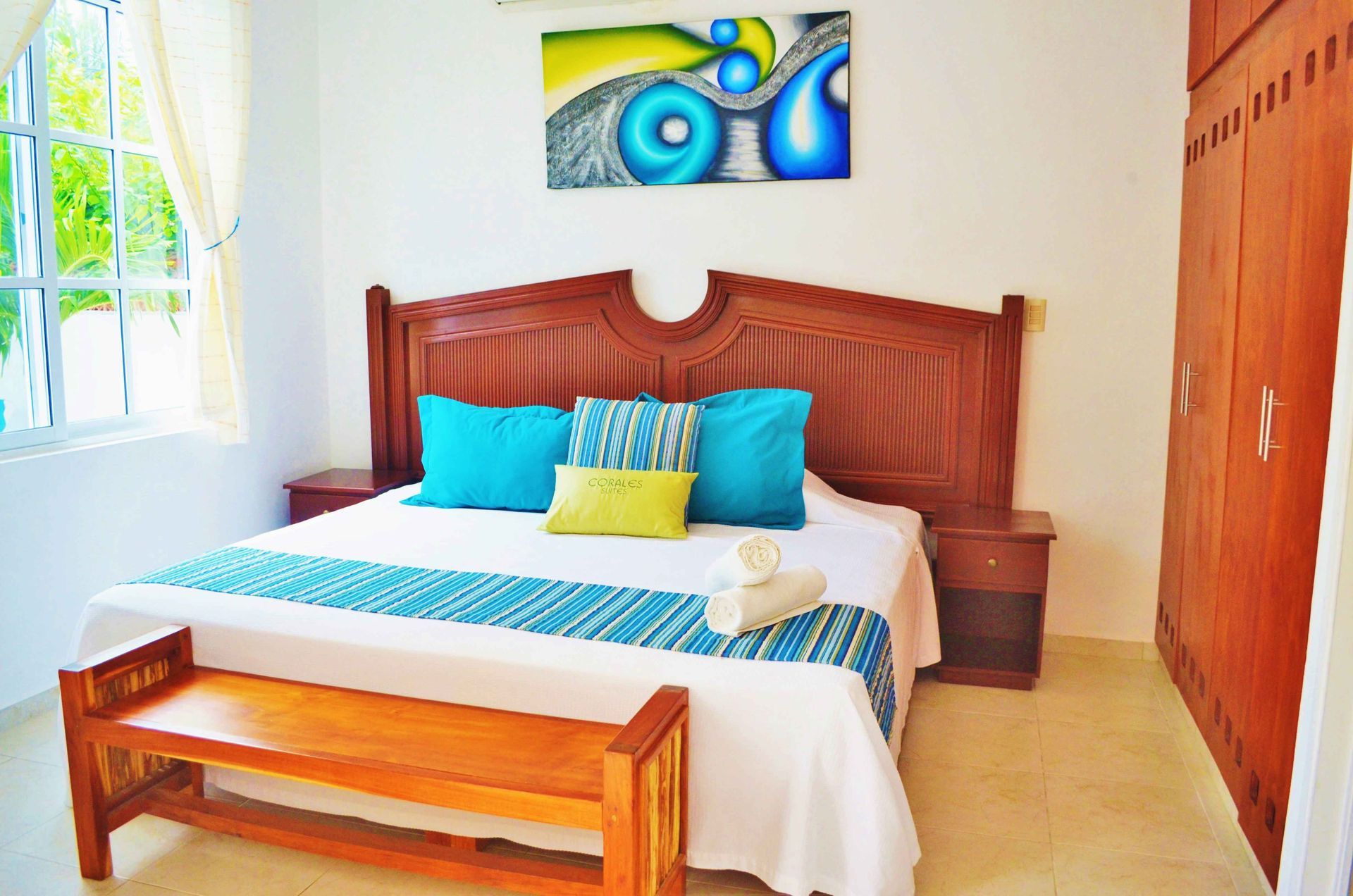 Dormitorio con un gran cabecero de madera, cama doble, banco y obras de arte, con detalles en azul y amarillo.