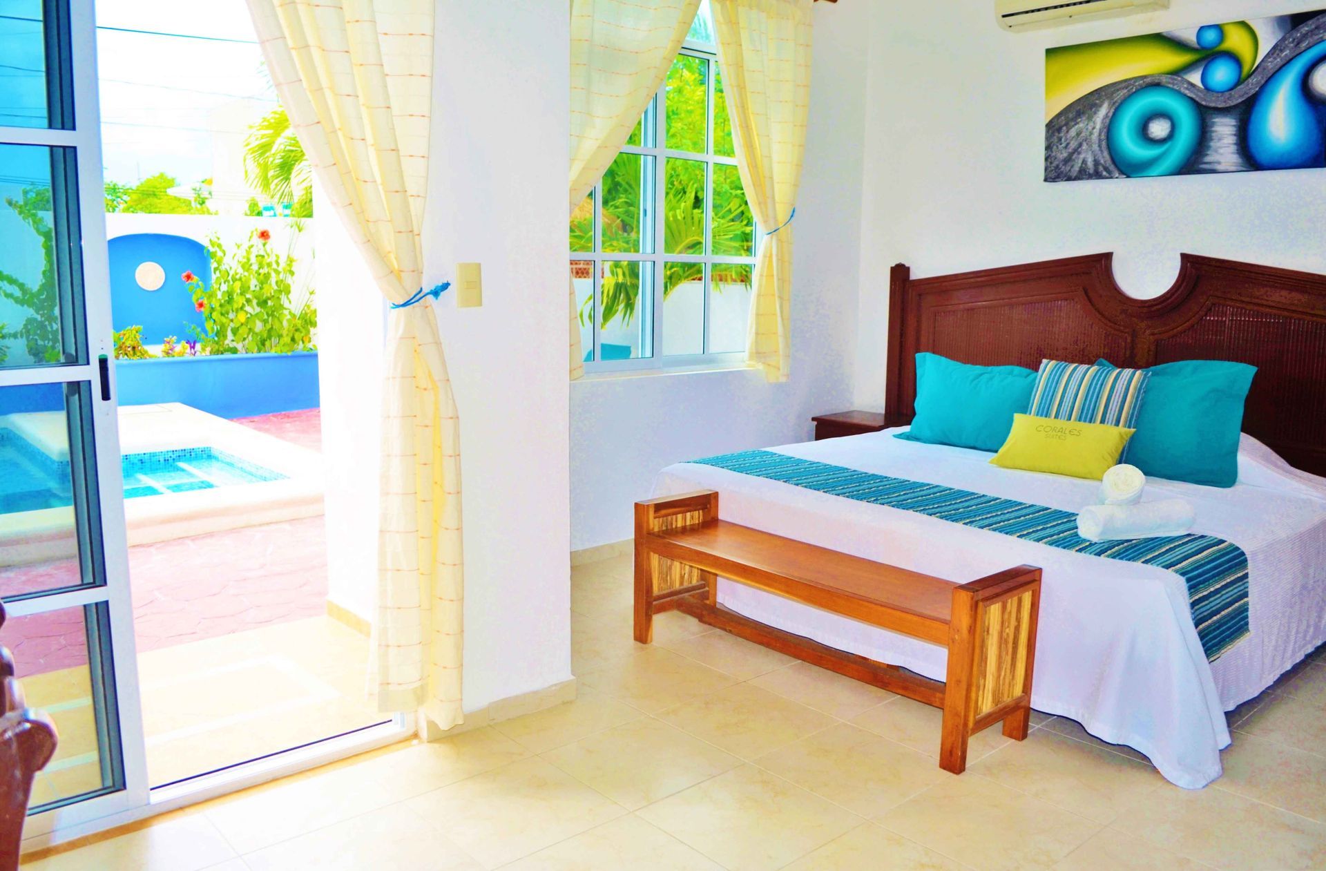 Dormitorio con vistas a la piscina; paredes blancas, detalles turquesa, muebles de madera, obras de arte.