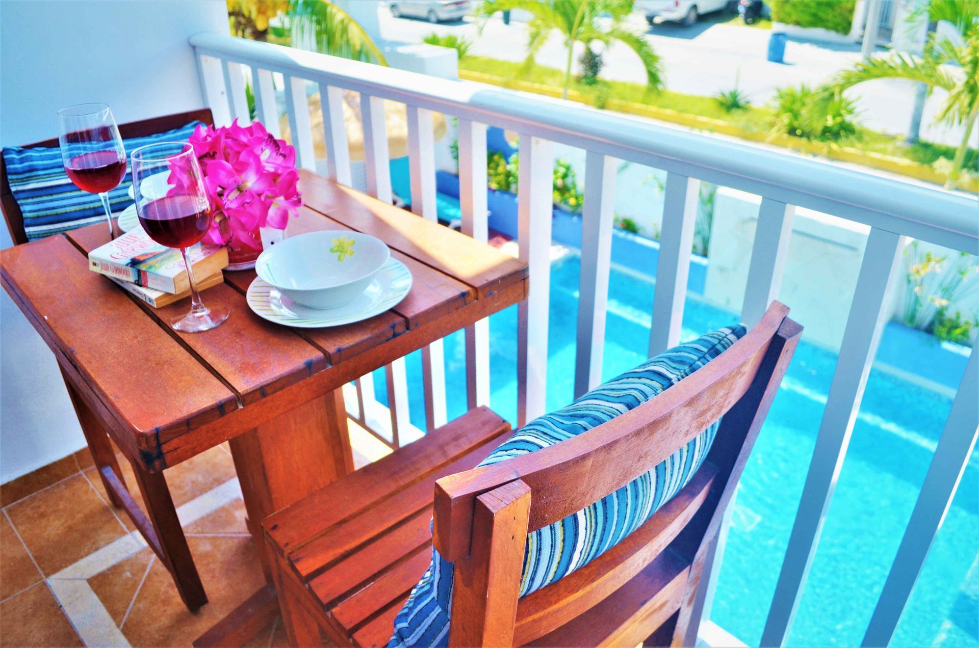 Small wooden balcony table set with wine, flowers, and a bowl, overlooking a sunny blue area with white railings.