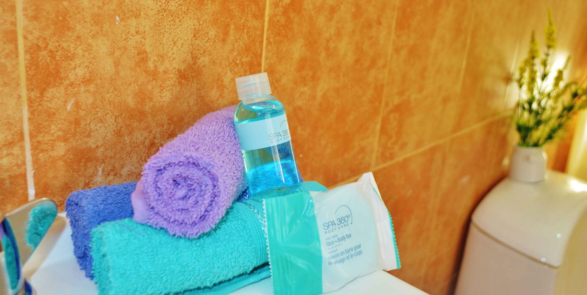 Toallas enrolladas, artículos de aseo y un jarrón con flores en un baño con azulejos naranjas.