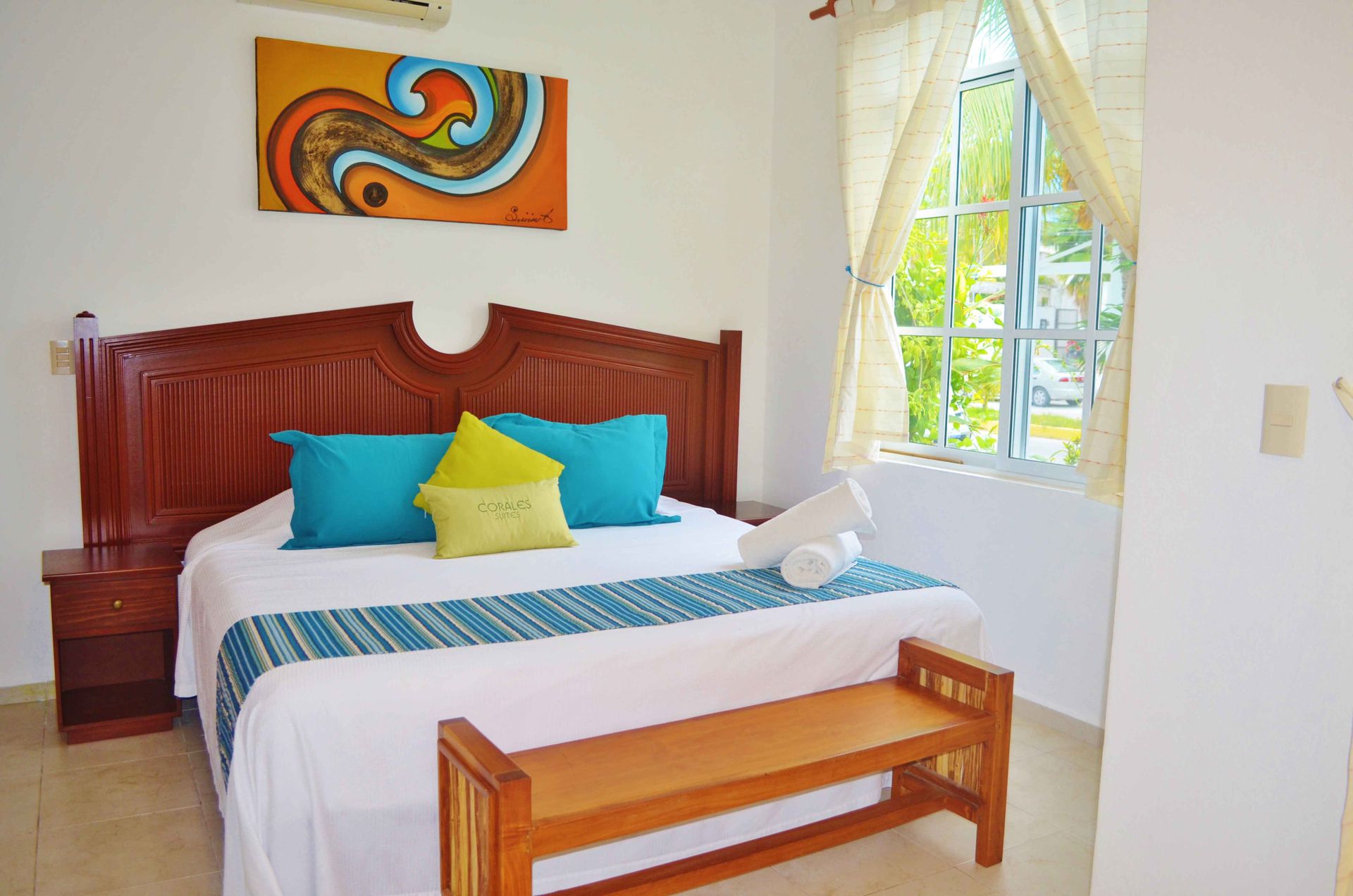 Dormitorio con cama, banco y obras de arte; paredes blancas y ventana.