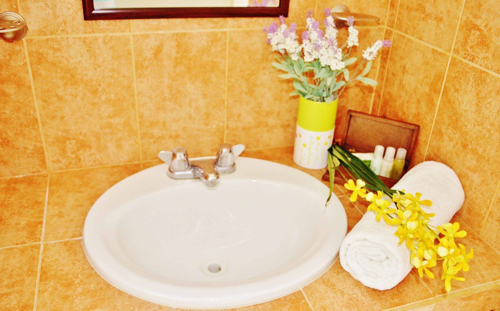 Lavabo blanco en un baño alicatado, con flores, toallas y artículos de aseo; combinación de colores marrón y amarillo.