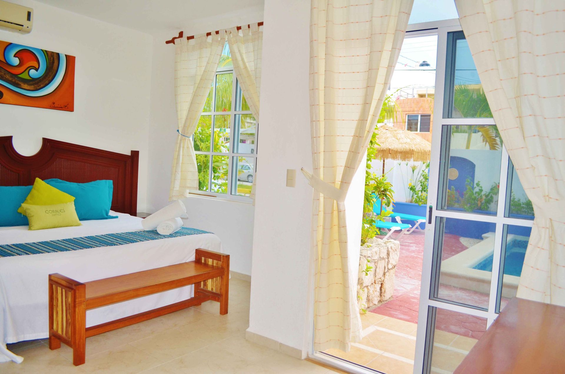 Dormitorio con puerta abierta a la zona de la piscina; cama, banco de madera, obras de arte, ventana y vistas a la piscina.