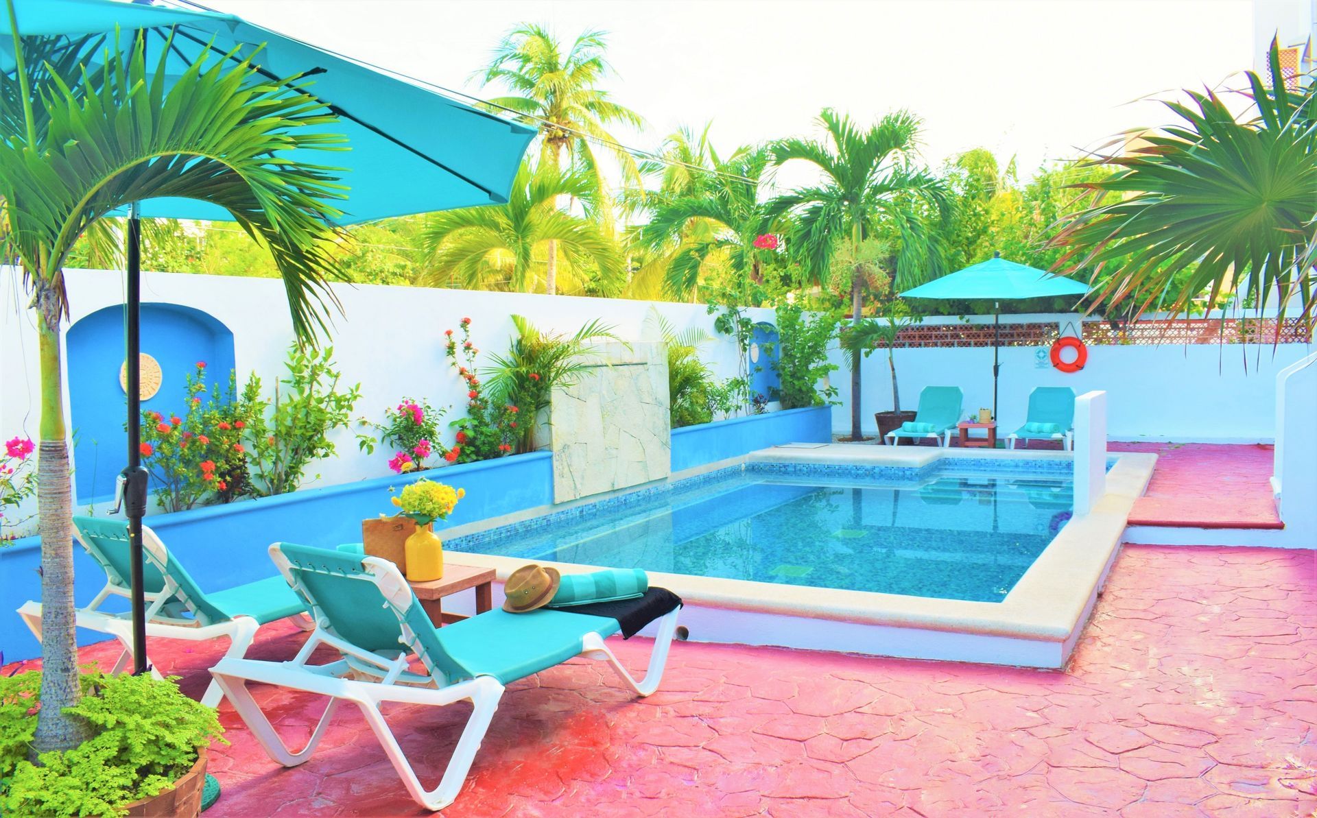 Pool with turquoise umbrellas, lounge chairs, and palm trees, surrounded by blue and white walls, pink patio.