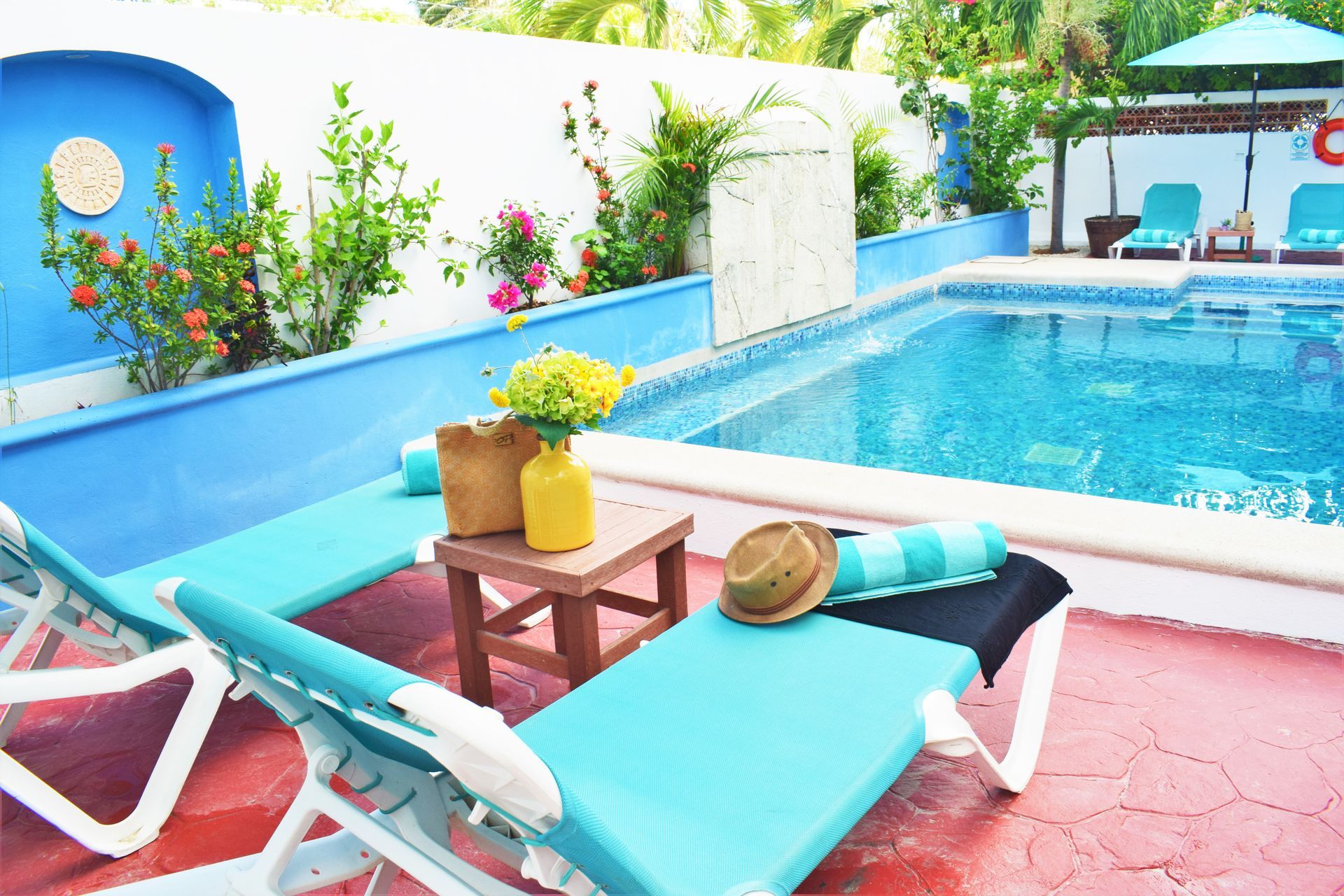 Poolside scene with turquoise lounge chairs, pool, and colorful flowers against a blue and white wall.