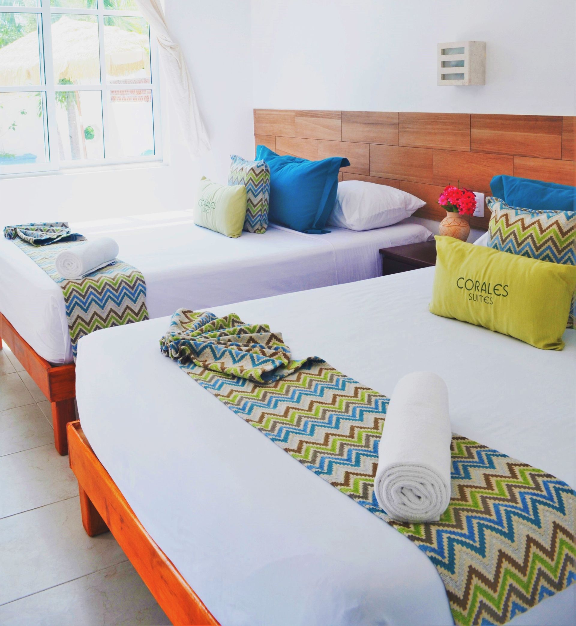 Dormitorio con dos camas, ropa de cama blanca, cabecero de madera, detalles en turquesa y amarillo, ventana al fondo.
