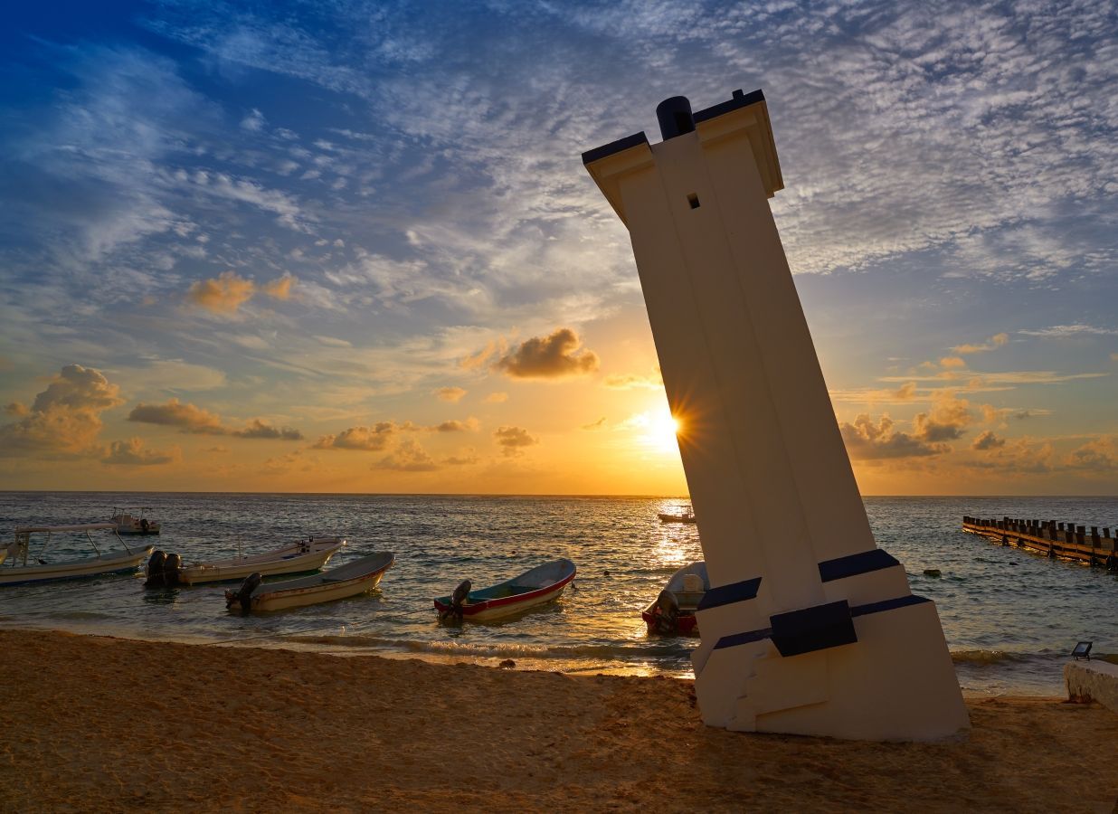 Leaning white lighthouse on beach, boats in water, sunset sky.