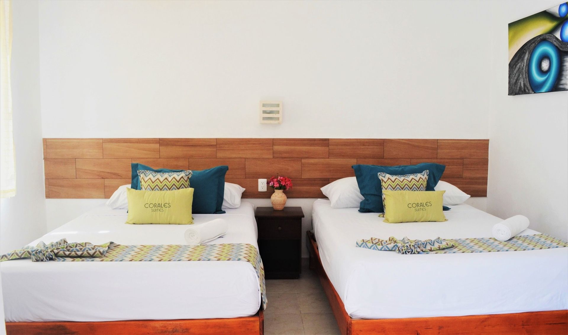 Dos camas con sábanas blancas, cabecero de madera, mesita de noche y cojines decorativos de colores en una habitación blanca.
