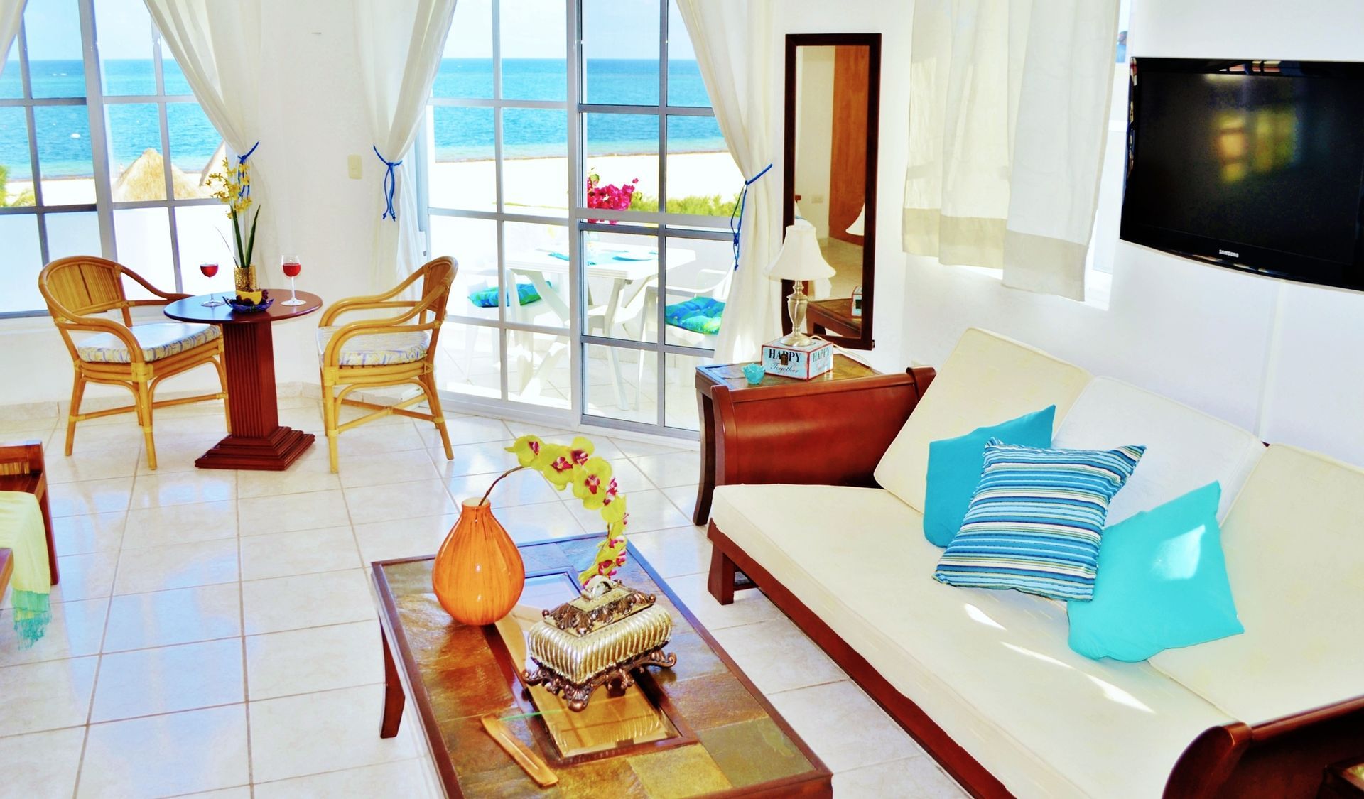 Sala de estar con vista al mar: muebles de mimbre, sofá blanco con cojines turquesa y televisor.