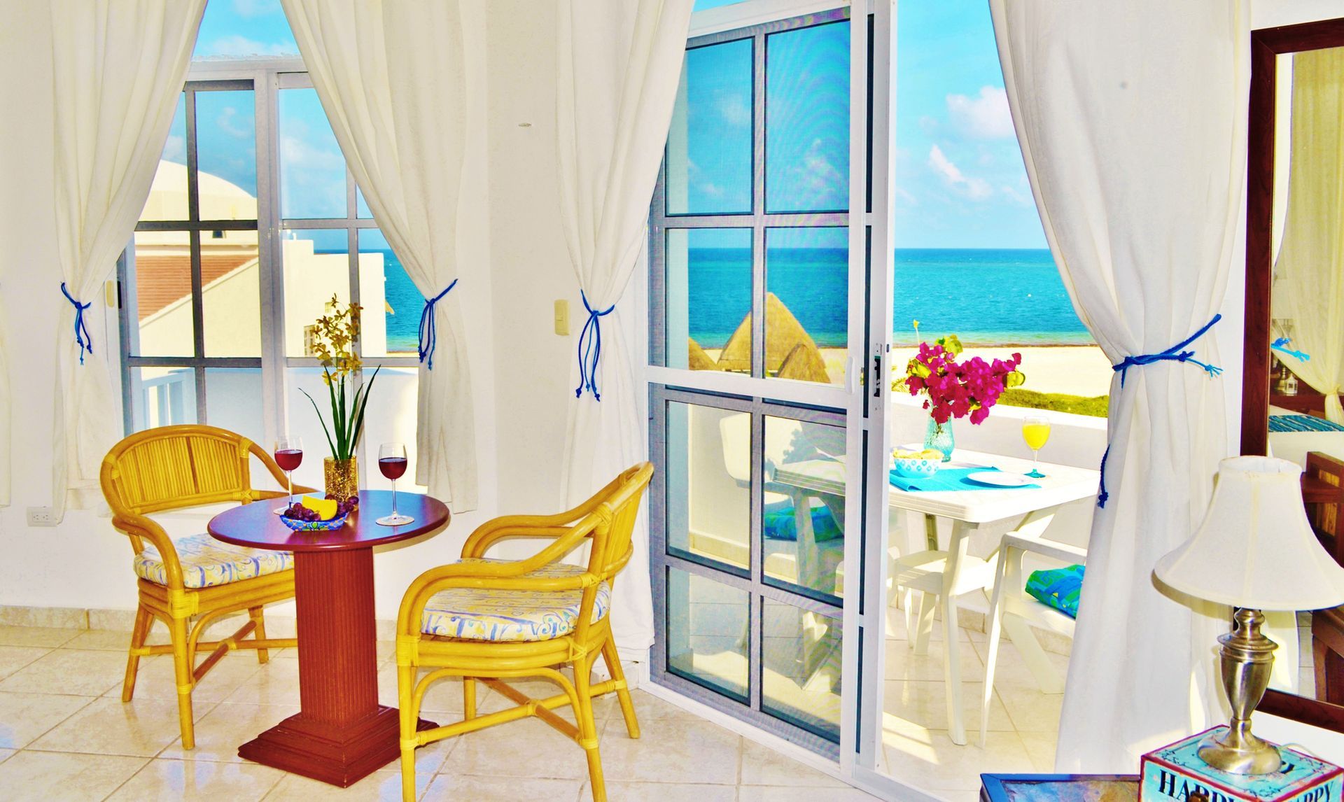 Vista interior de un balcón con vistas al océano, con mesa, sillas y cortinas blancas.