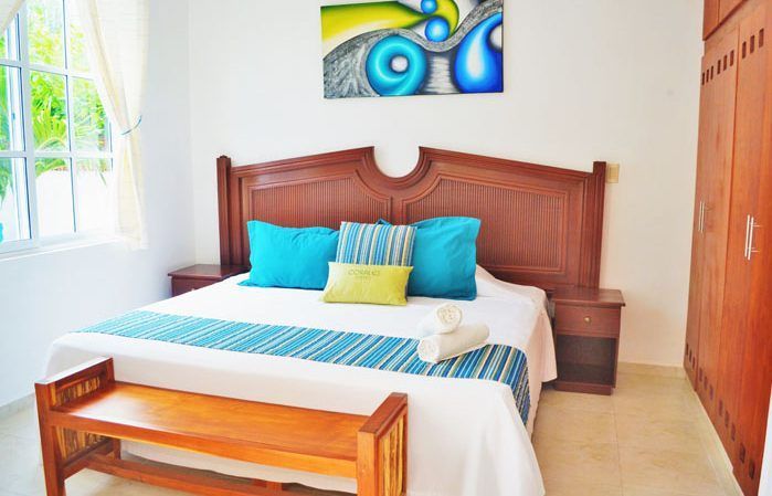 Dormitorio con cama grande, almohadas coloridas, muebles de madera y obras de arte abstractas.