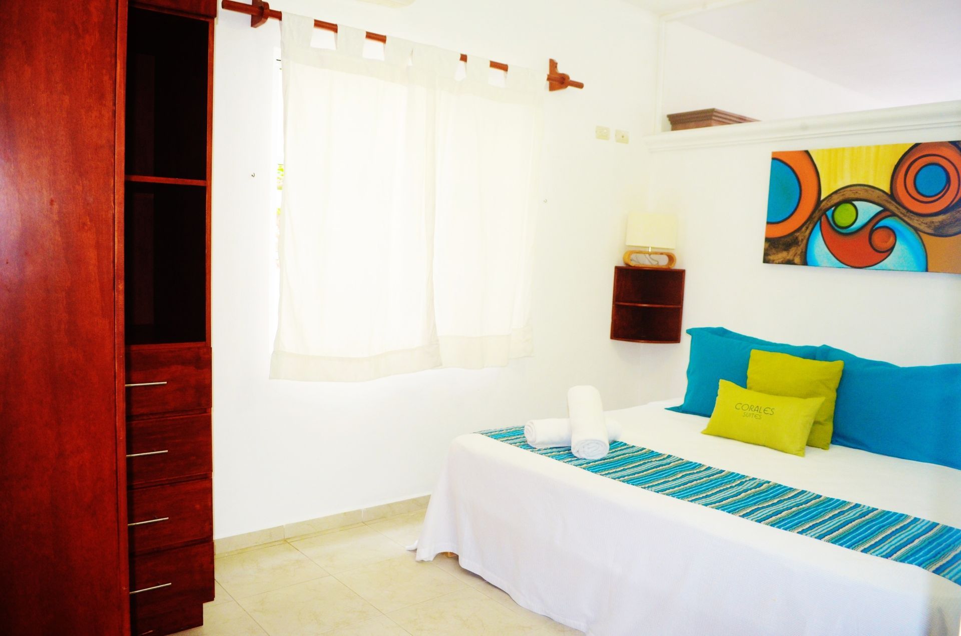 Dormitorio con paredes blancas, armario de madera, cama con detalles turquesa y obras de arte coloridas.