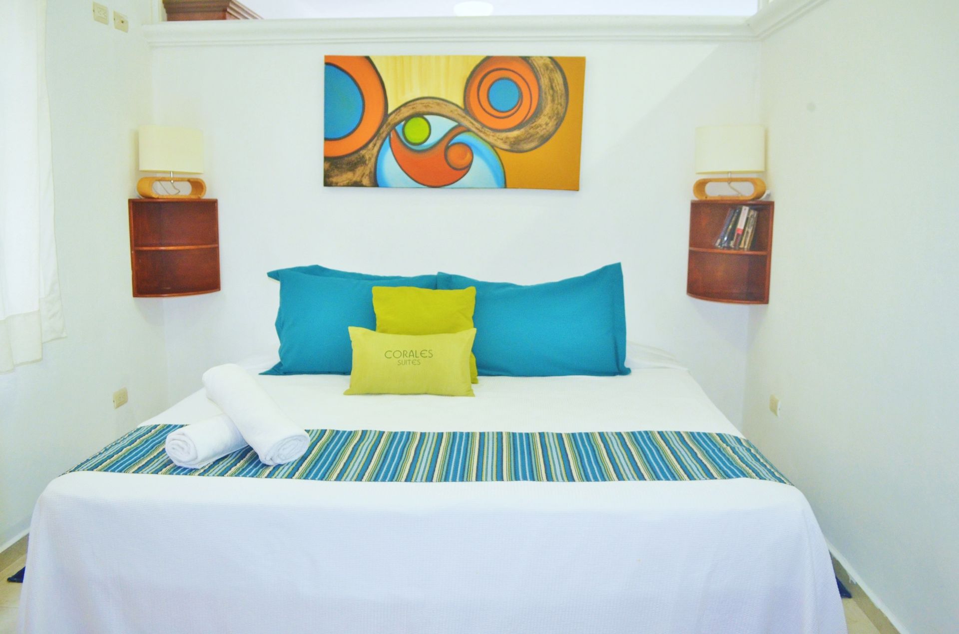 Dormitorio con cama blanca, almohadas coloridas, obras de arte y mesillas de noche de madera.