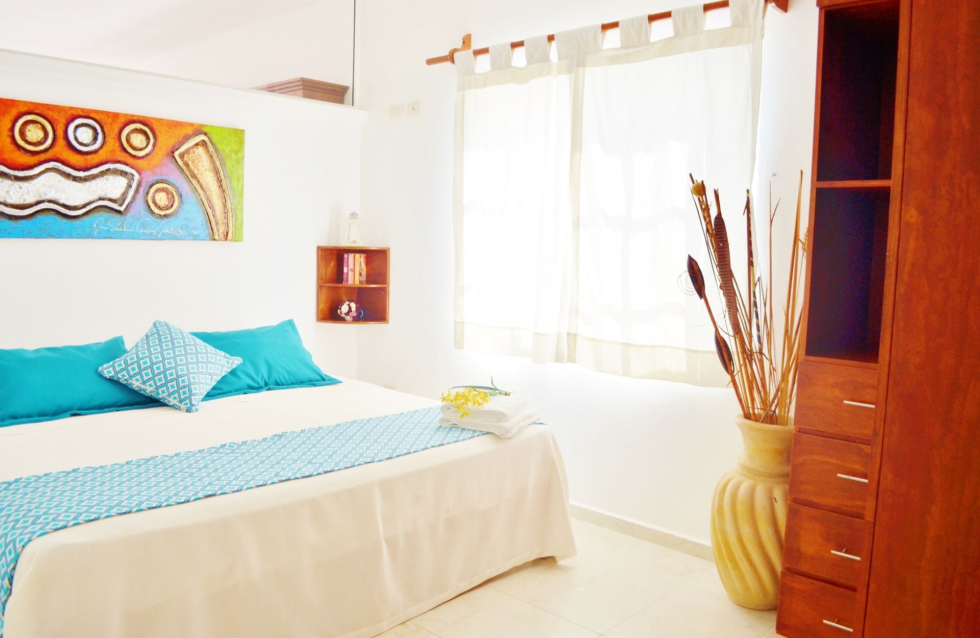 Dormitorio con cama, decoración azul y blanca, obras de arte, jarrón alto, armario de madera y ventana luminosa.