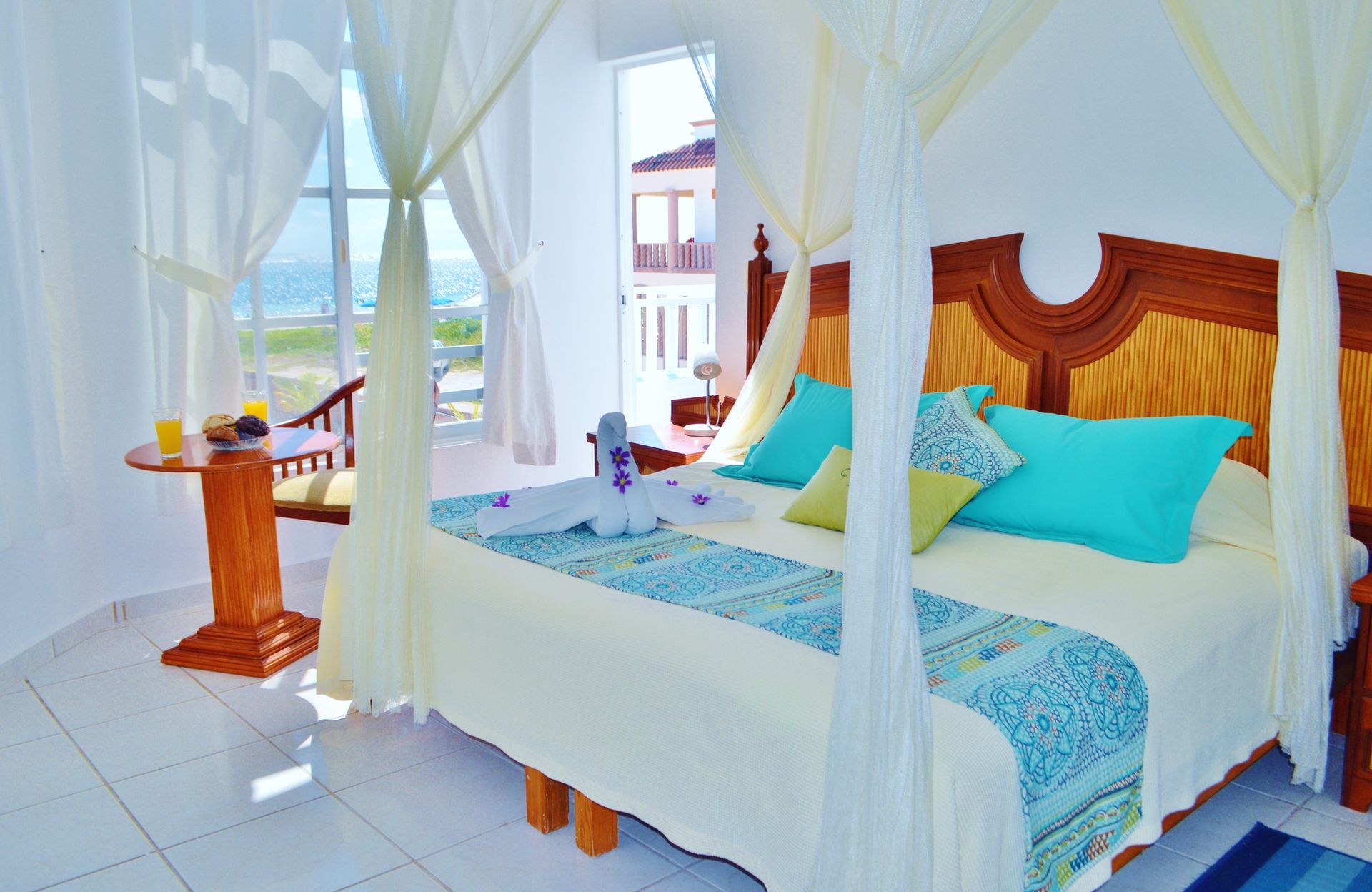 Dormitorio con cama con dosel, vista al mar, decoración en tonos verde azulado y blanco, mesa con desayuno.