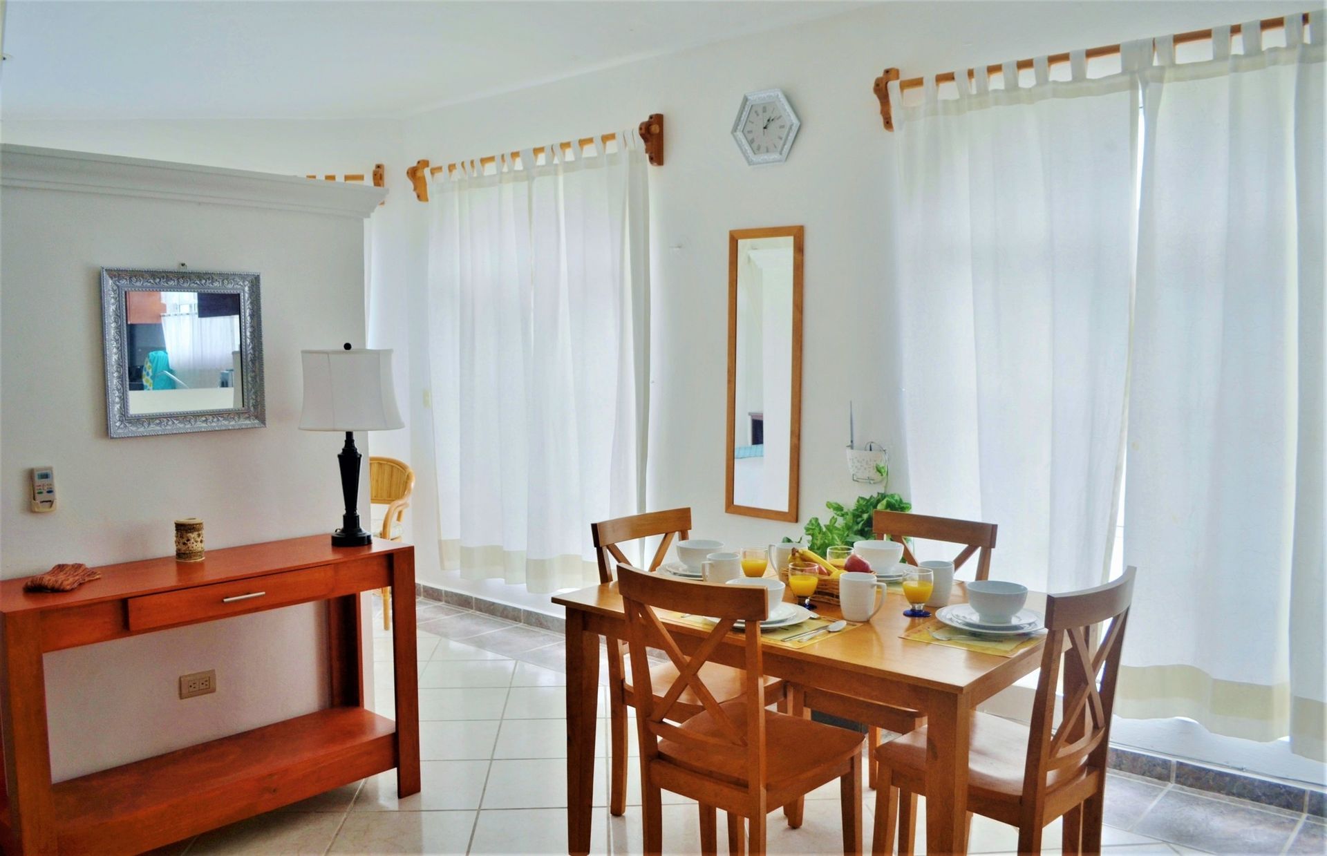 Dining room with wooden table set with breakfast, white curtains, and a console table.
