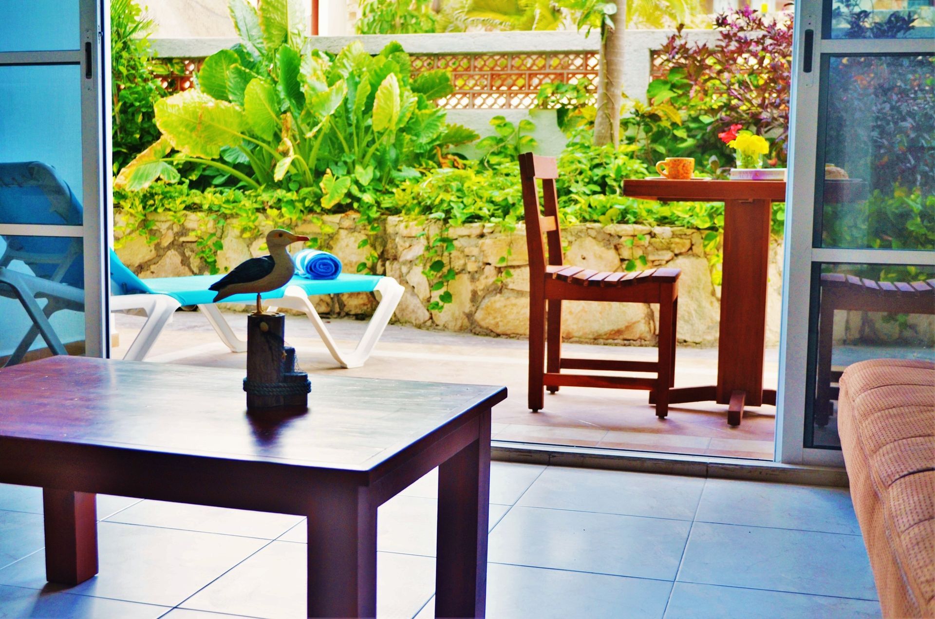 Interior view with open doors to a patio. Dark wood furniture, lush green plants, and a sun lounger are visible.