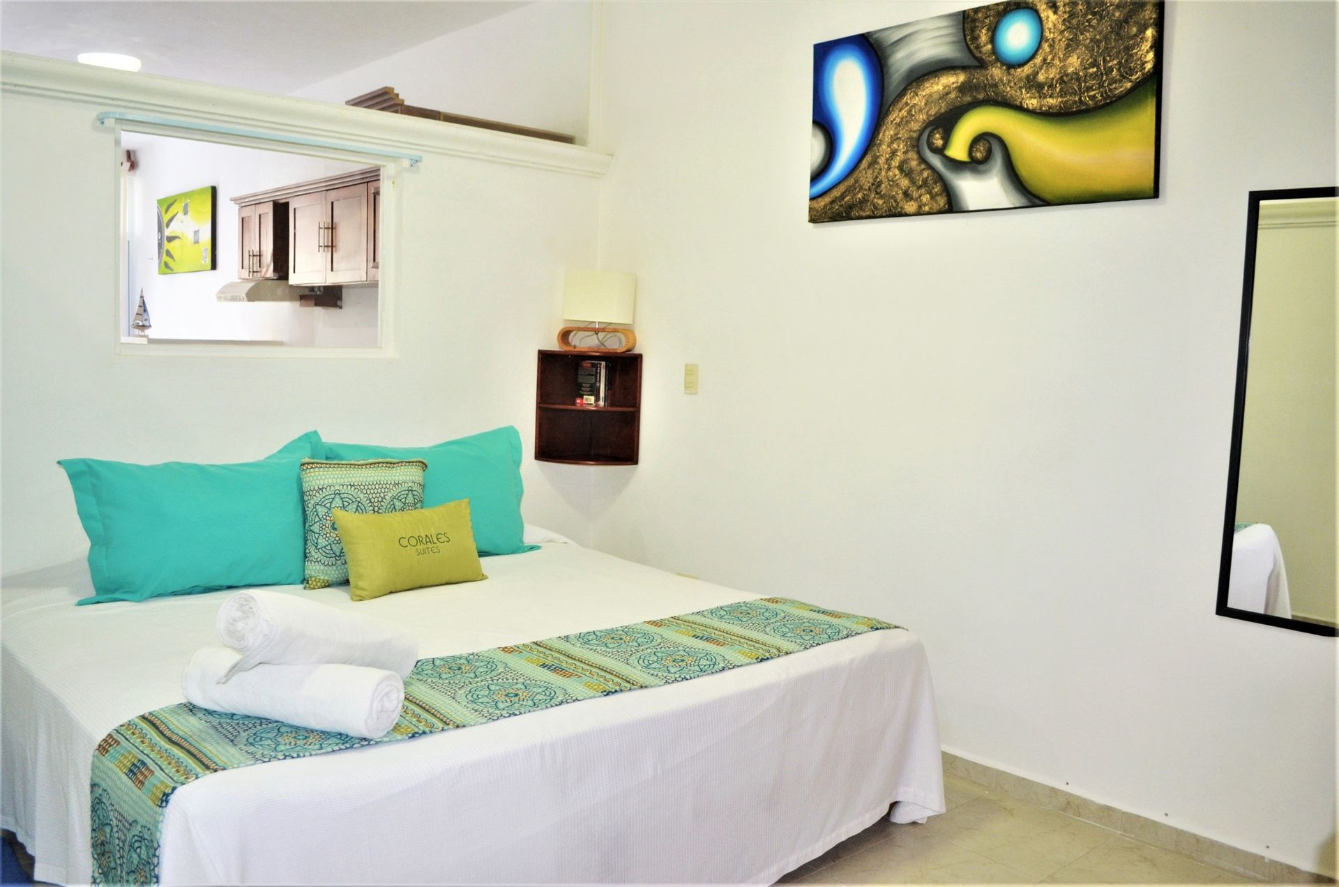 Bedroom with white walls, a bed with teal pillows, artwork, and a small kitchen visible through a window.