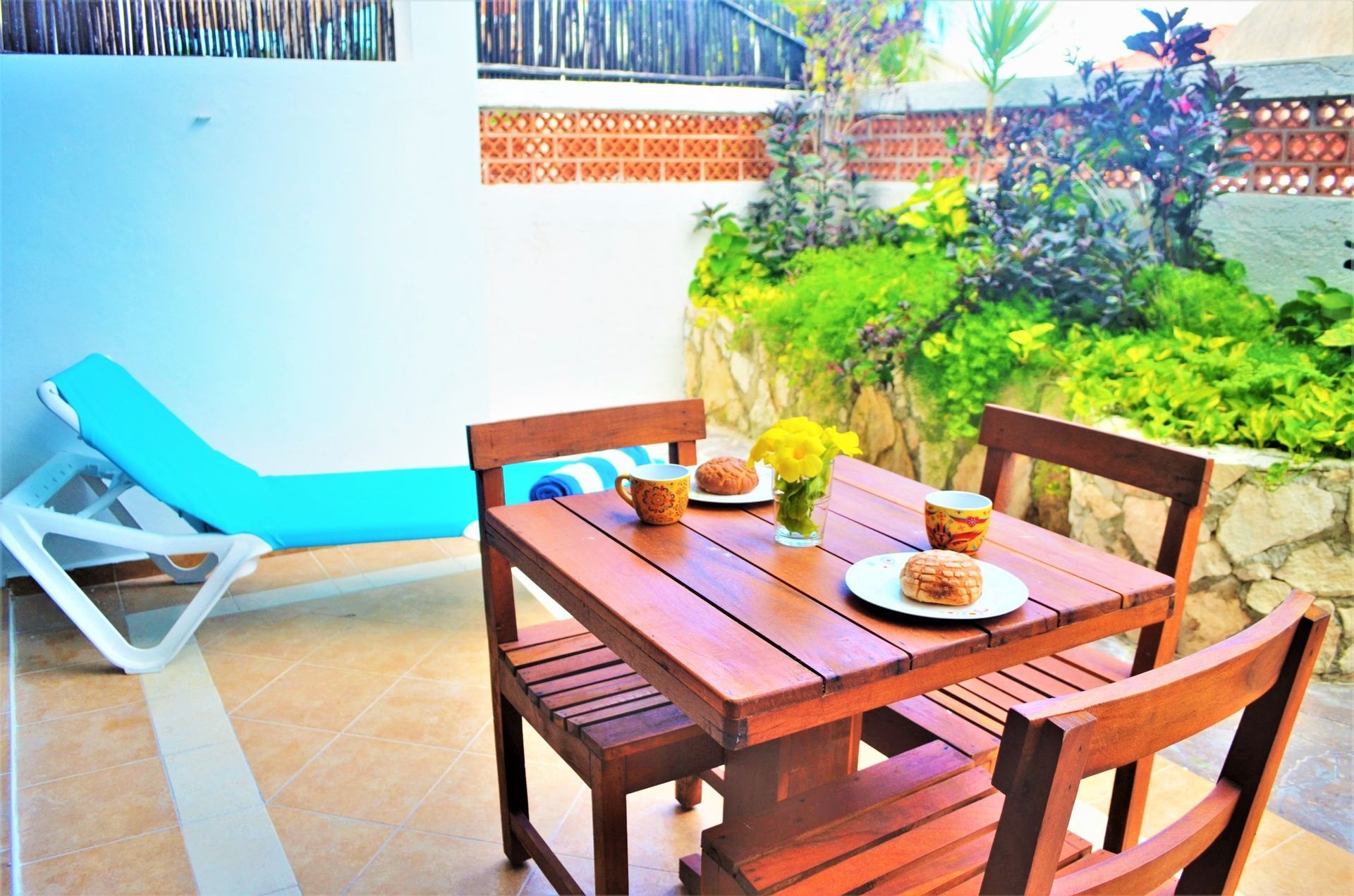 Outdoor patio with wooden table set for two, lounge chair, and garden.