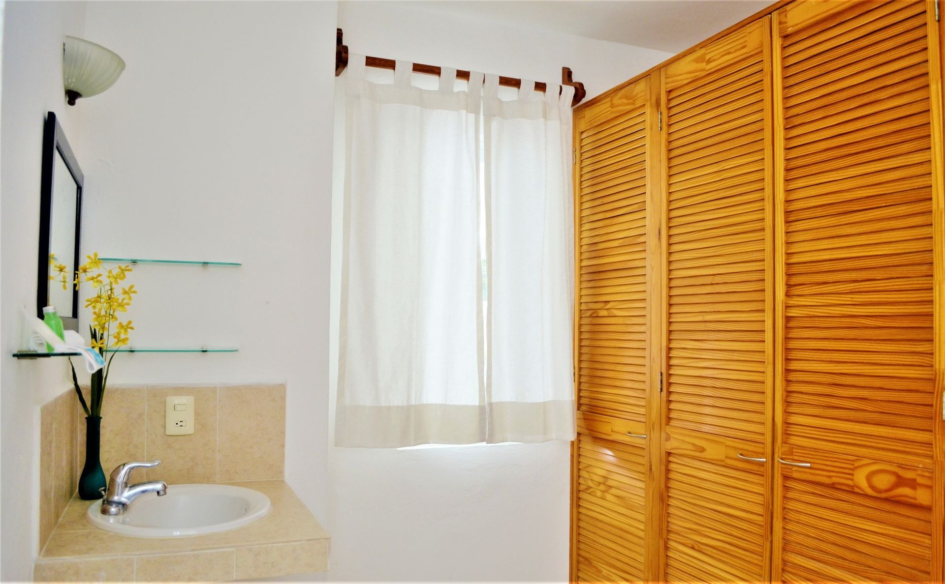Bathroom with white walls, a vanity, window with sheer curtain, and wooden closet doors.