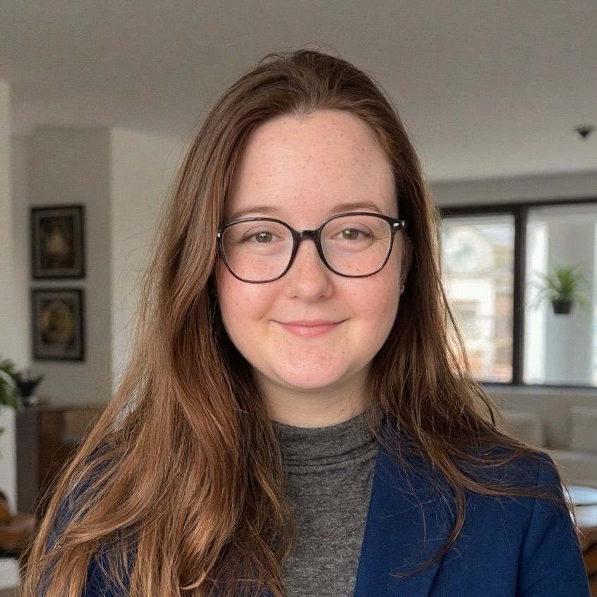 Person with glasses and long brown hair smiling in a home interior, wearing a blue blazer.