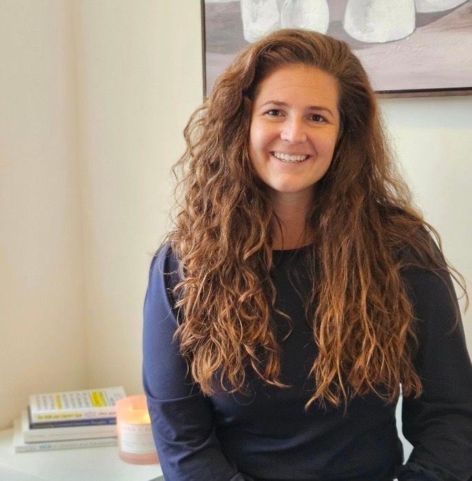 Smiling person with long curly hair in a dark top, seated indoors against a light wall.