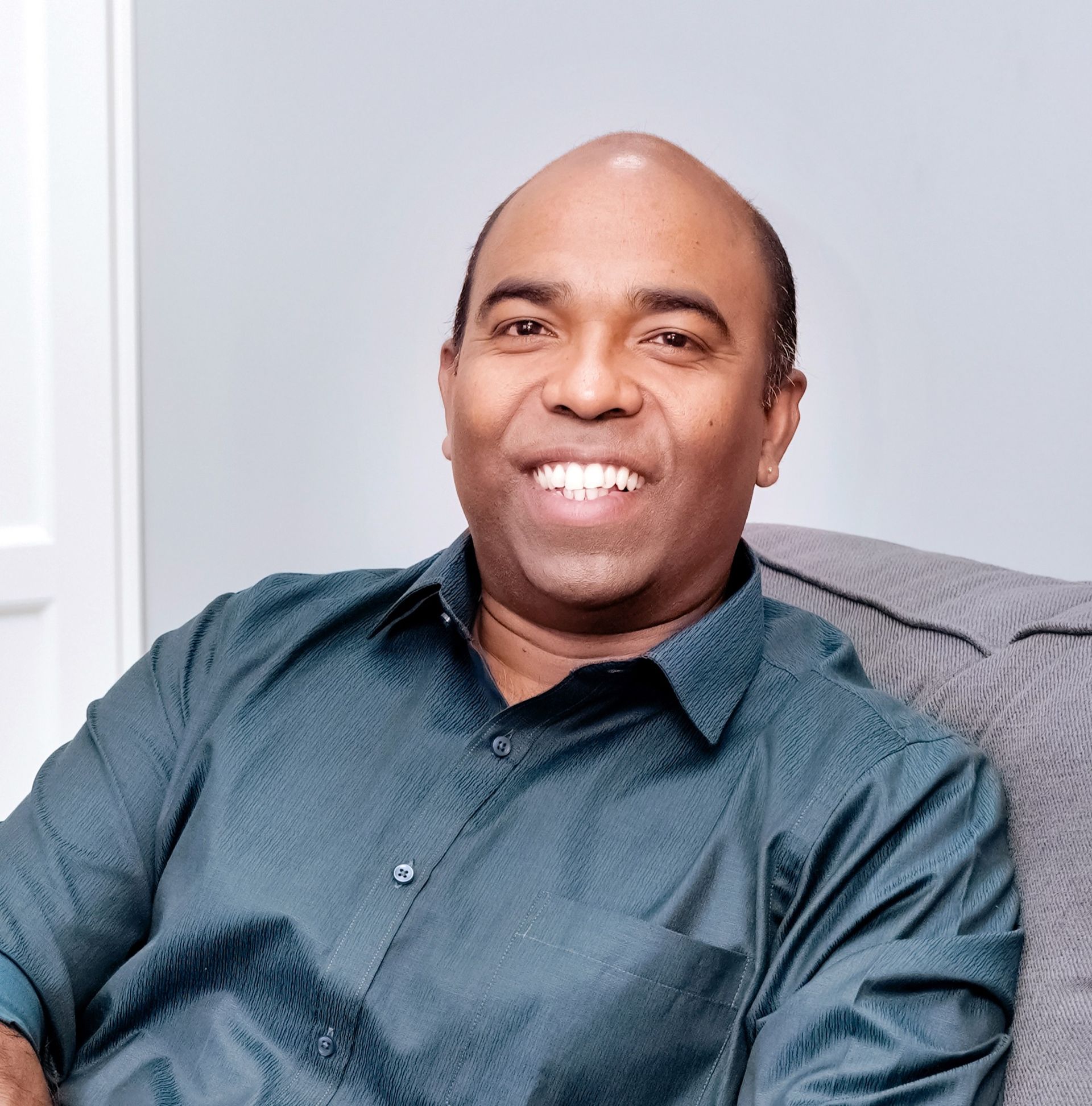 Smiling man in a teal shirt seated on a gray couch indoors