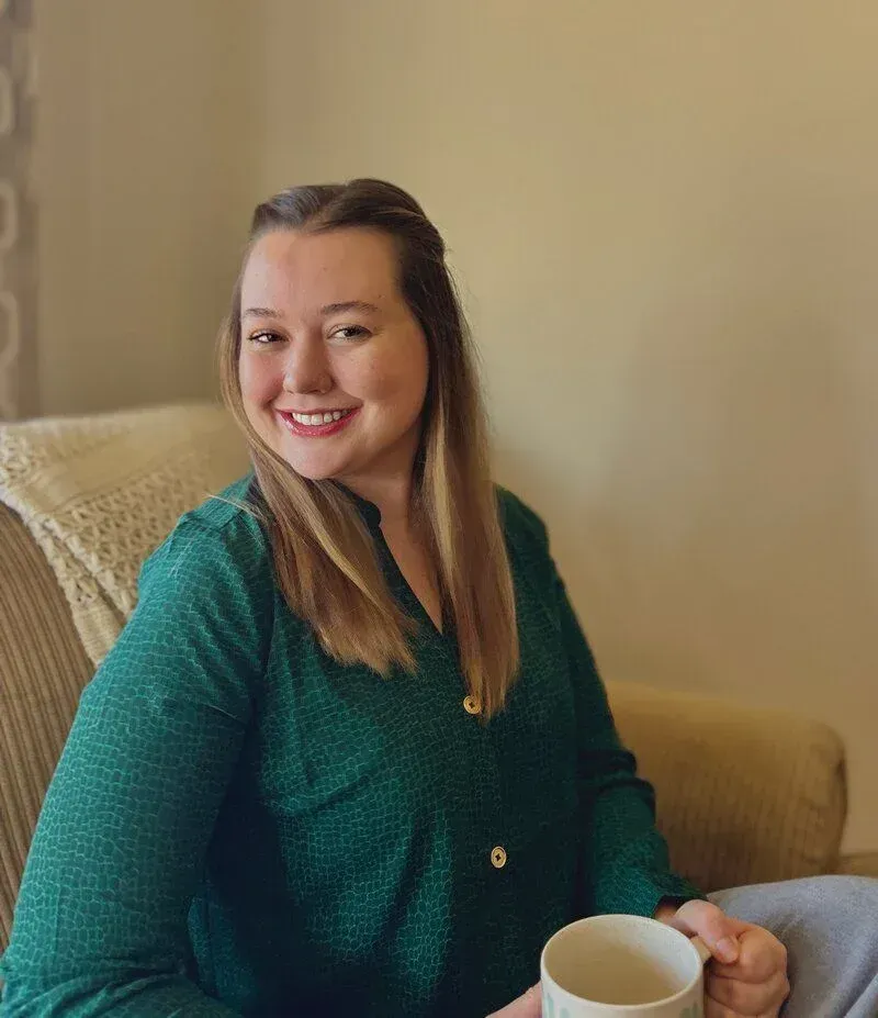Smiling woman in a green sweater seated on a beige couch, holding a white mug.