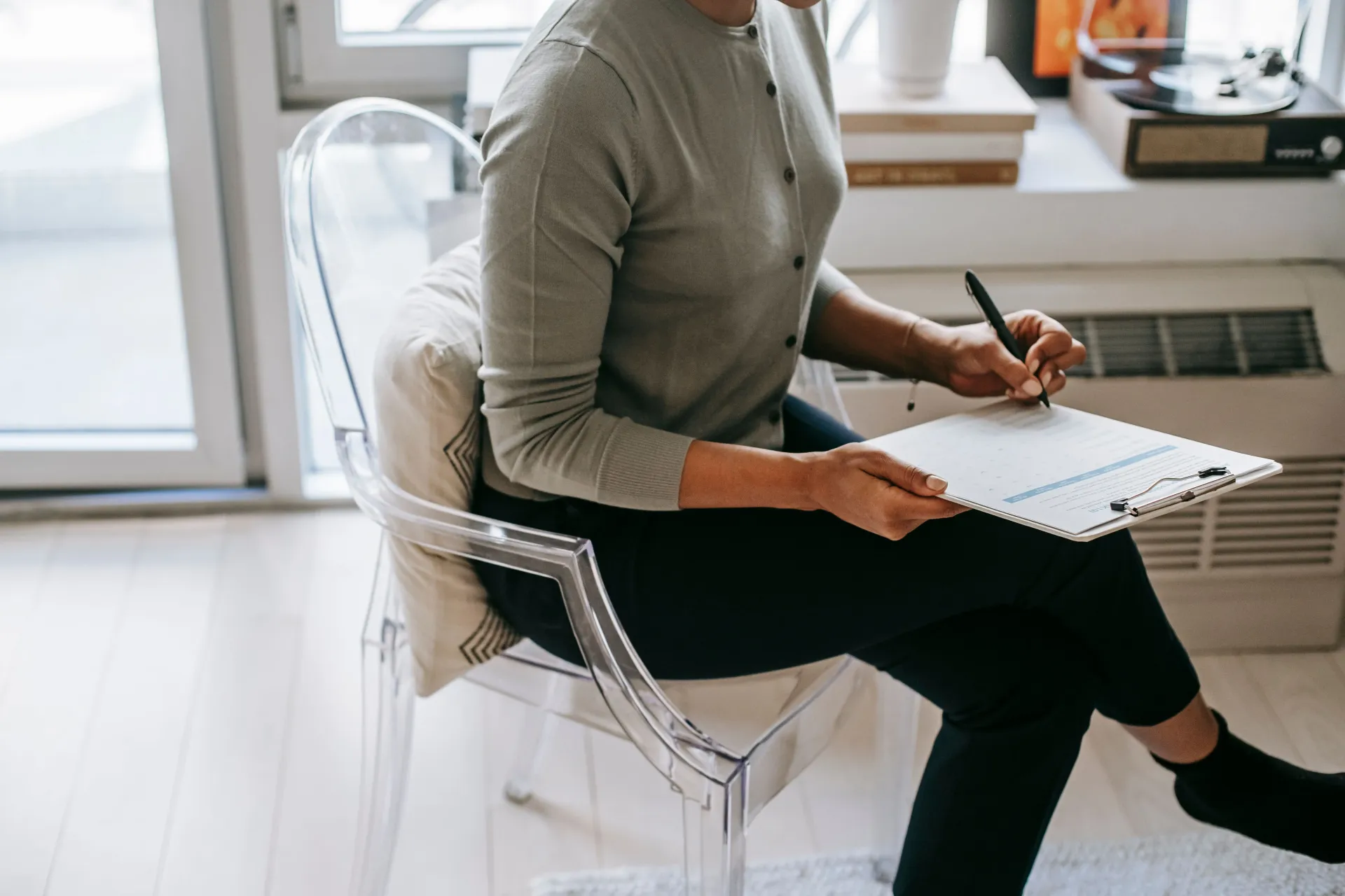 Person seated in a clear chair, writing in a notebook near a bright window